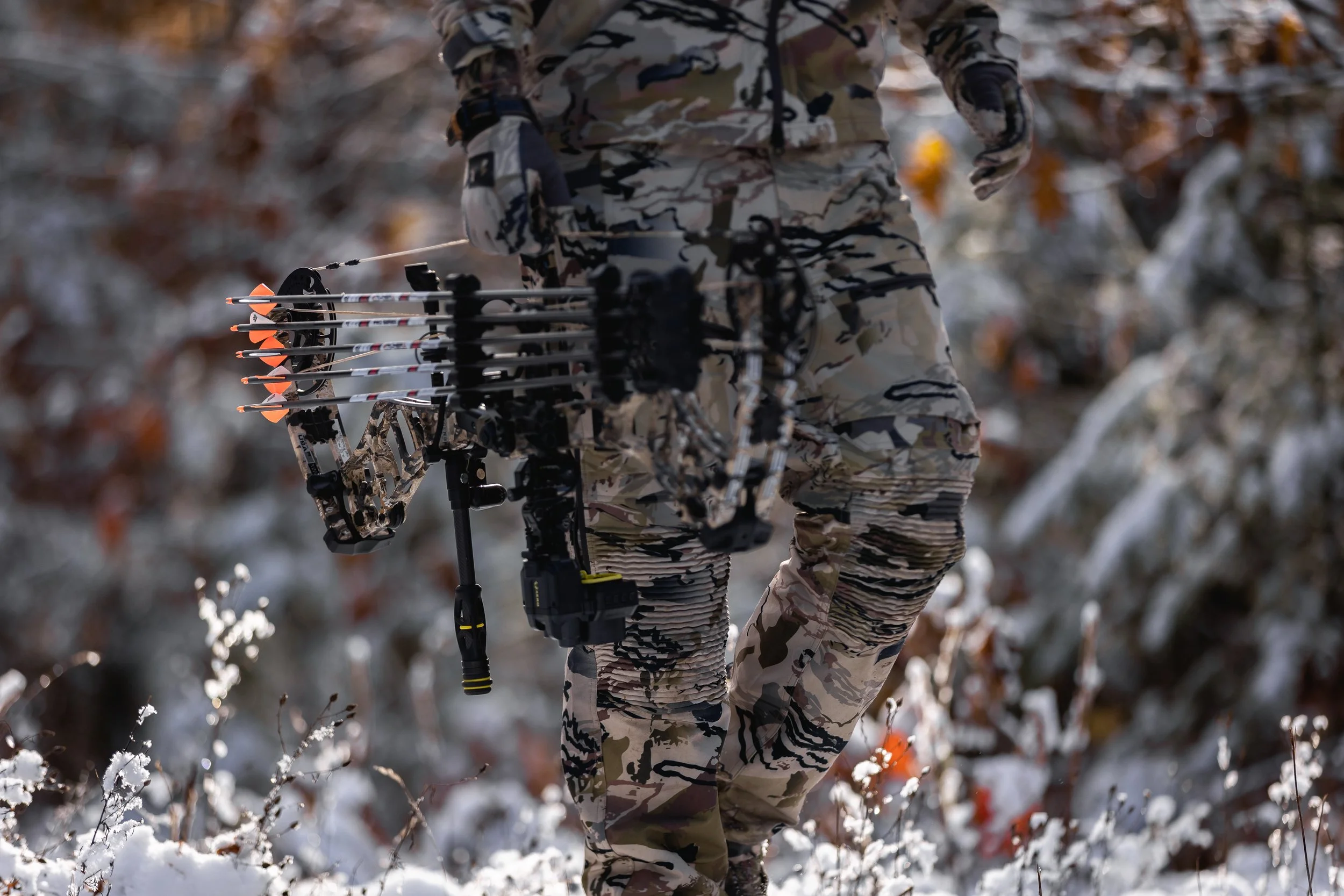 Person in camouflage clothing holding a compound bow with arrows, standing in a snowy outdoor setting with blurred trees and snow-covered foliage in the background.