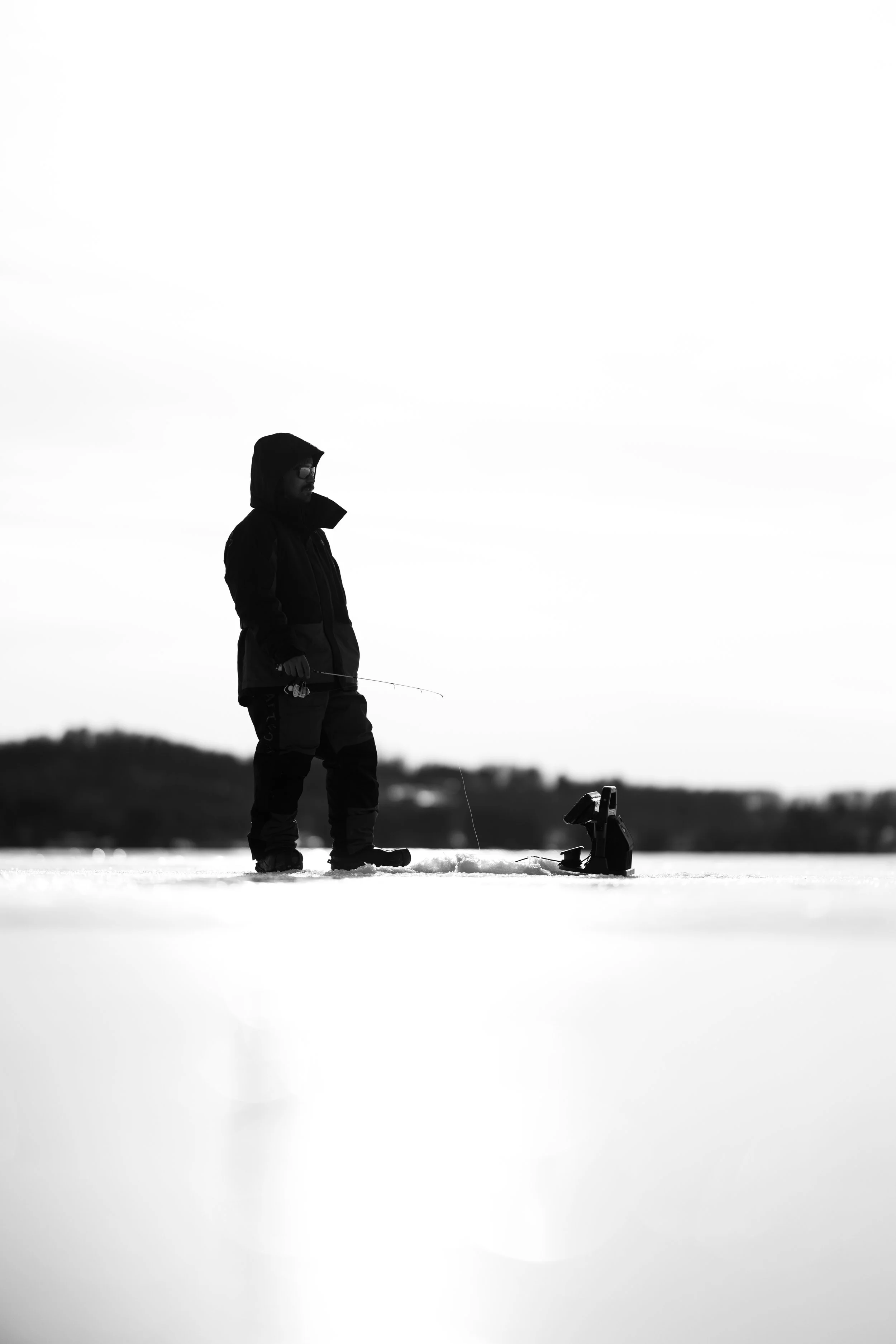 Silhouette of a person ice fishing on a snow-covered landscape with a fishing rod and a small ice fishing shelter.