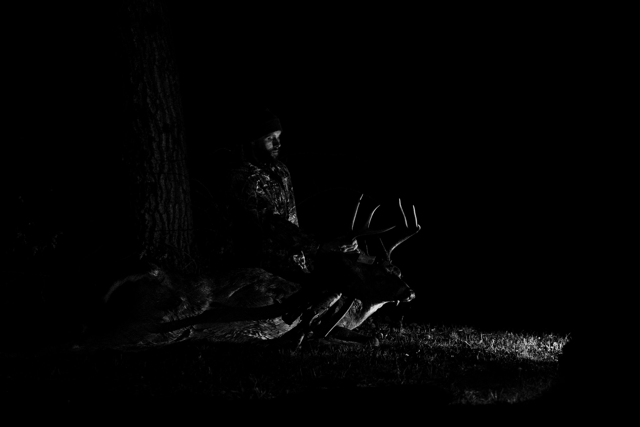 A person sitting on the ground near a tree at night, illuminated by a light source, possibly holding a camera or another device.
