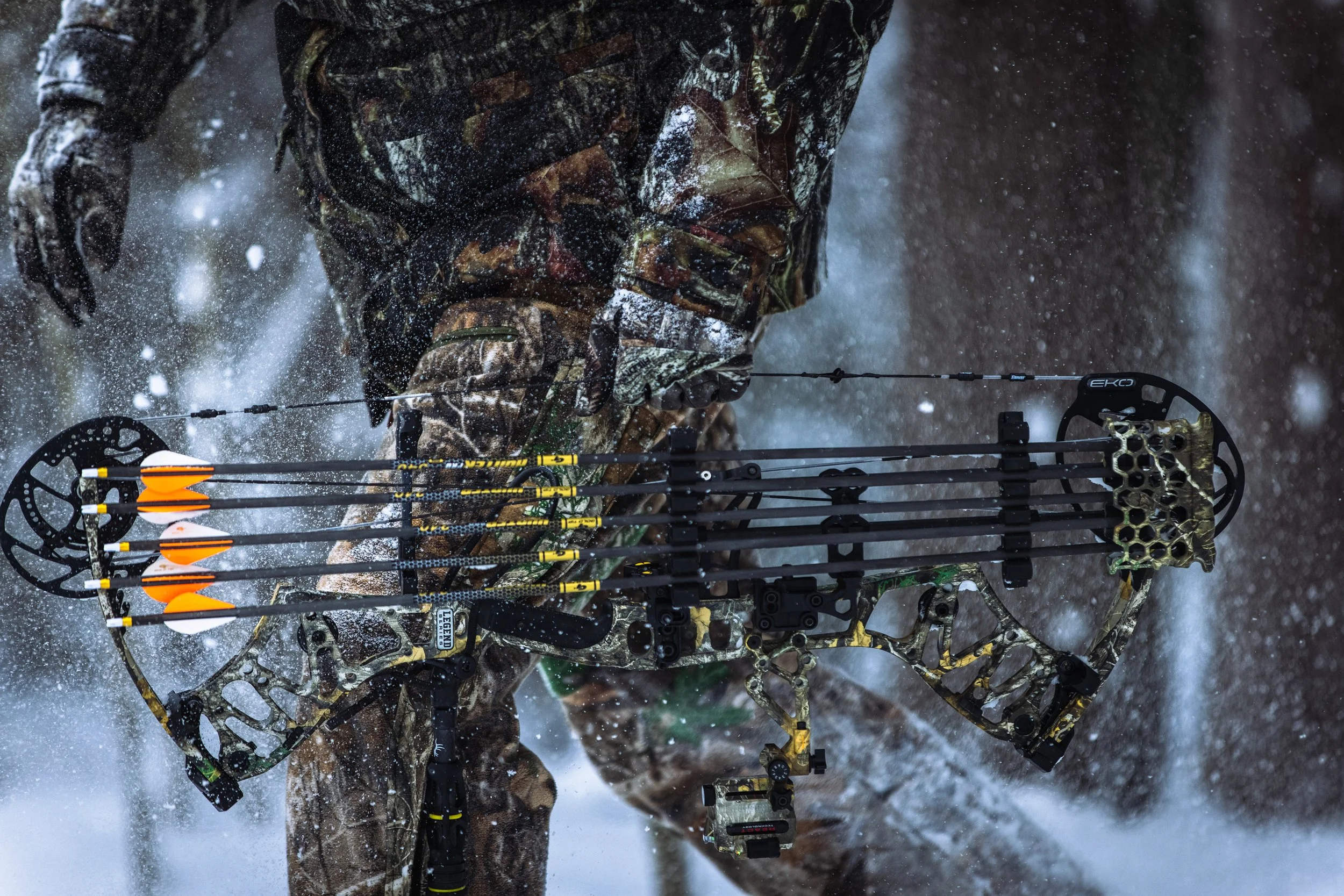 Close-up of a person in camouflage clothing holding a compound bow with arrows, shooting through snow.