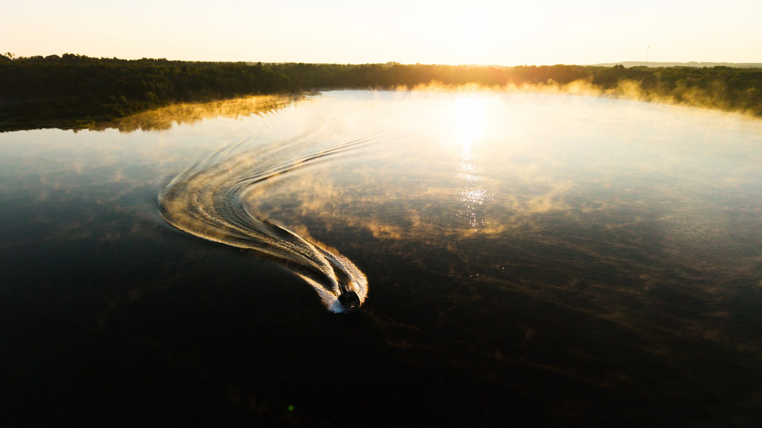 A boat moving across a calm body of water during sunrise or sunset, creating ripples and reflecting the sky and surrounding landscape.
