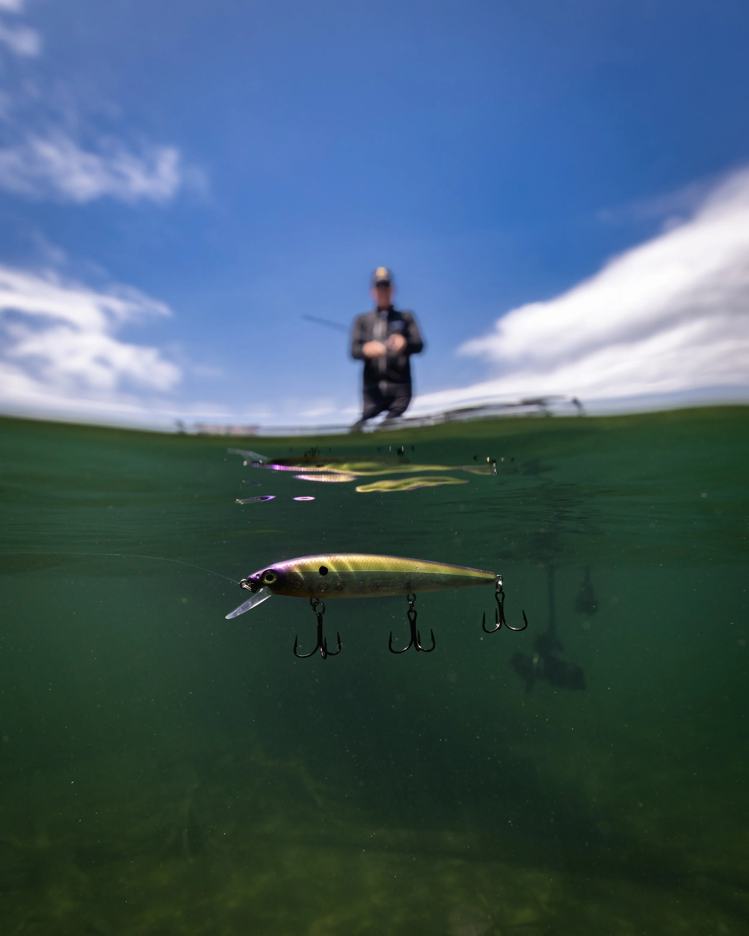 Underwater view of a fishing lure with fish hooks, with a person holding a fishing rod blurred in the background against a blue sky.