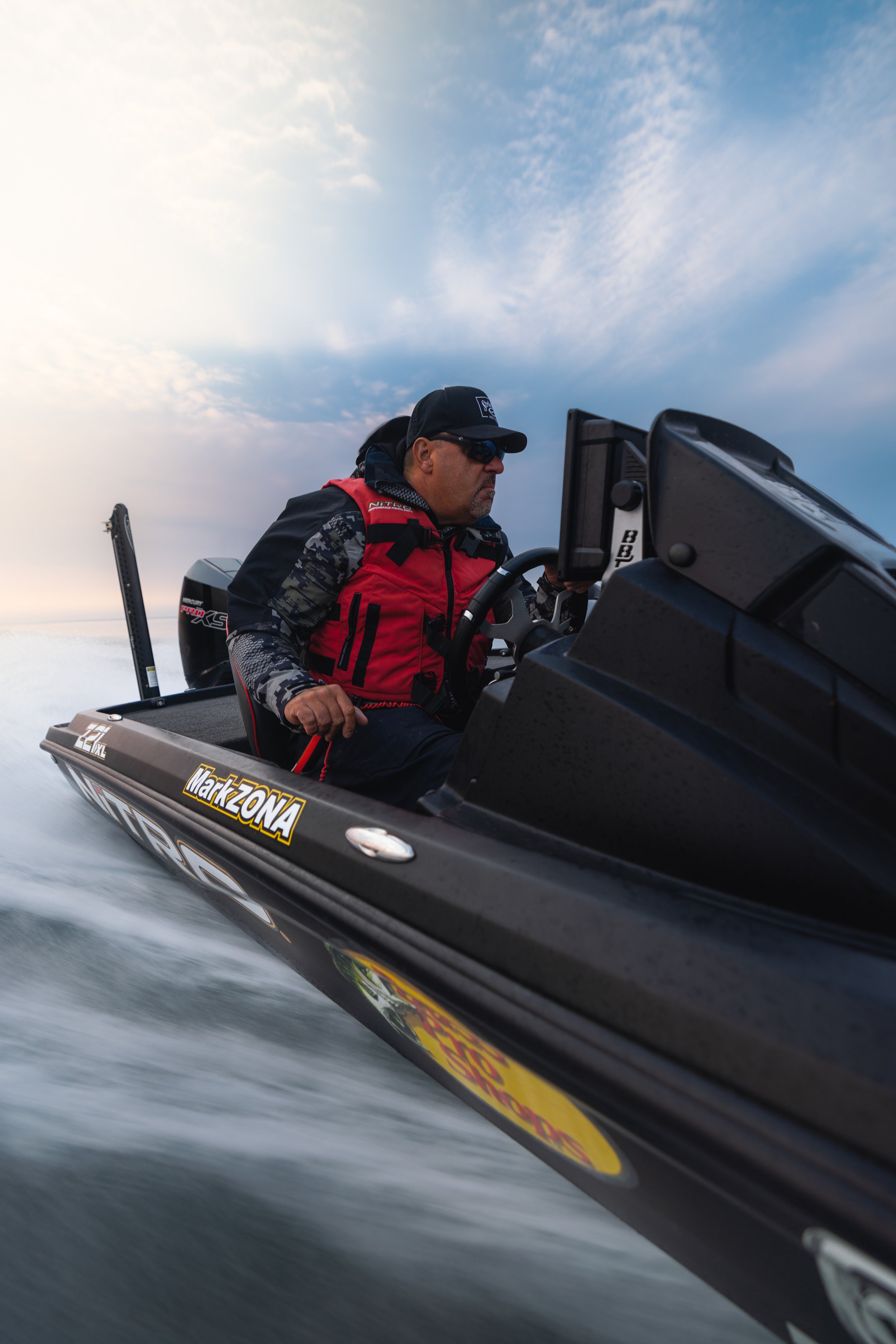Man in a red and black life vest operating a black boat with stickers, on water under a cloudy sky.