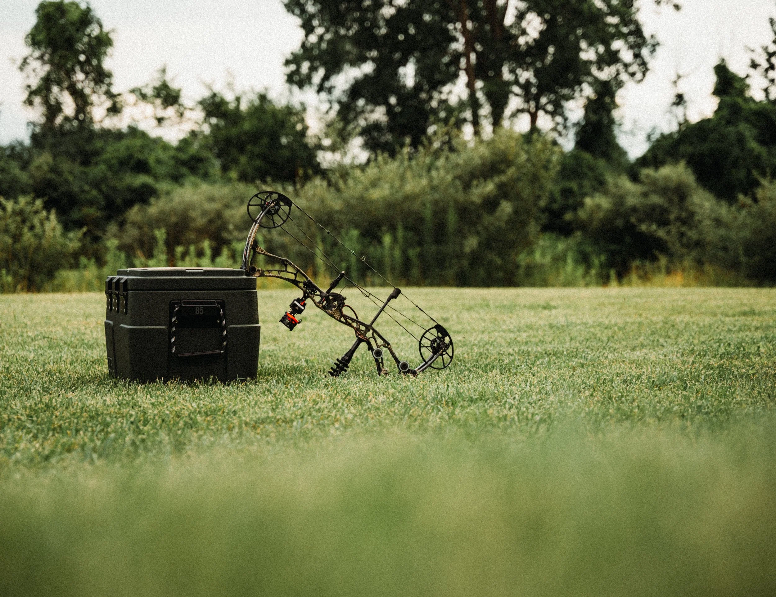 A compound bow leaning against a black storage container on a grassy field with trees in the background.