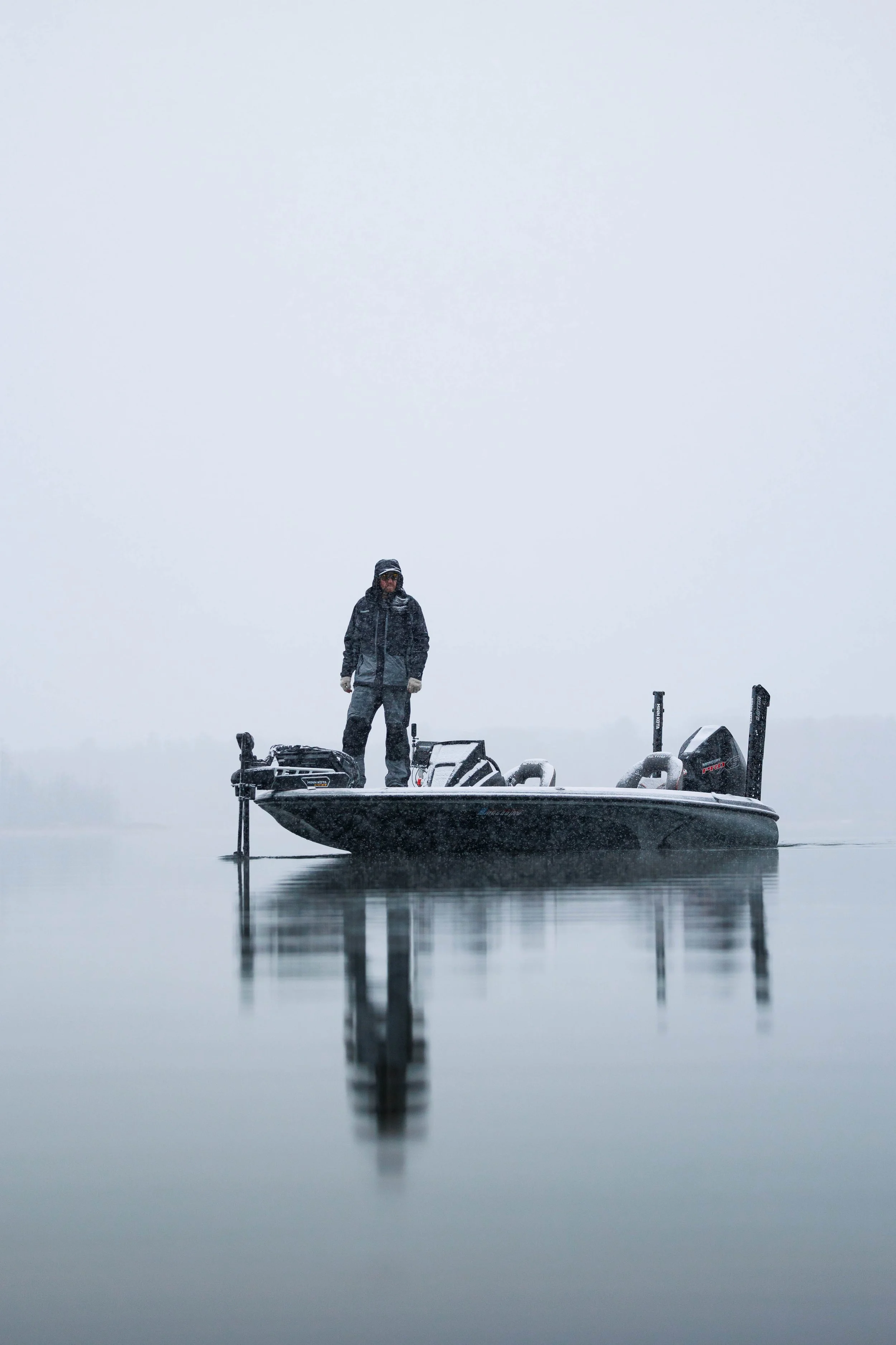 A person dressed in winter clothing standing on a small fishing boat in a foggy, calm body of water.