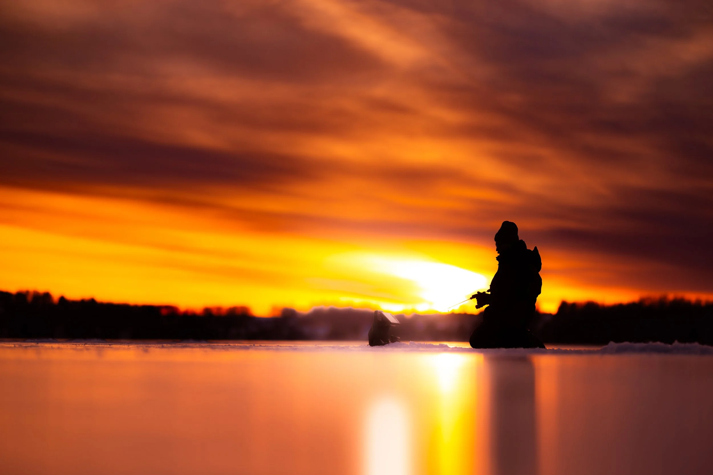 Silhouette of a person ice fishing at sunset with colorful sky and reflected light on the ice
