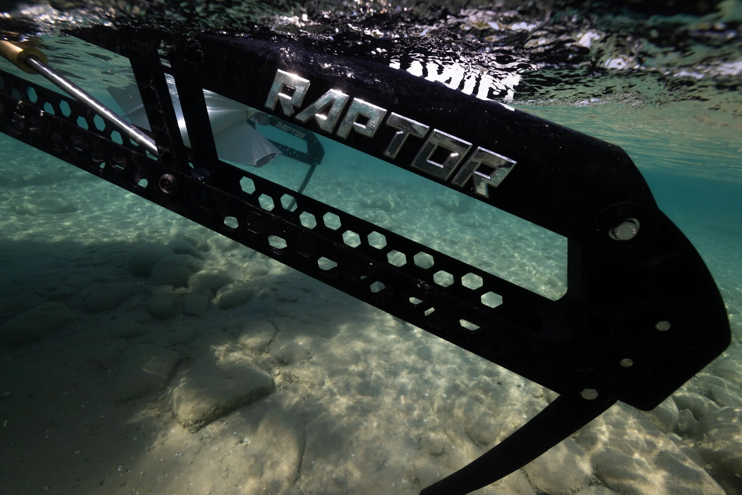 Underwater view of a black Raptor paddleboard with silver Raptor logo, partially submerged in clear water with a rocky bottom.
