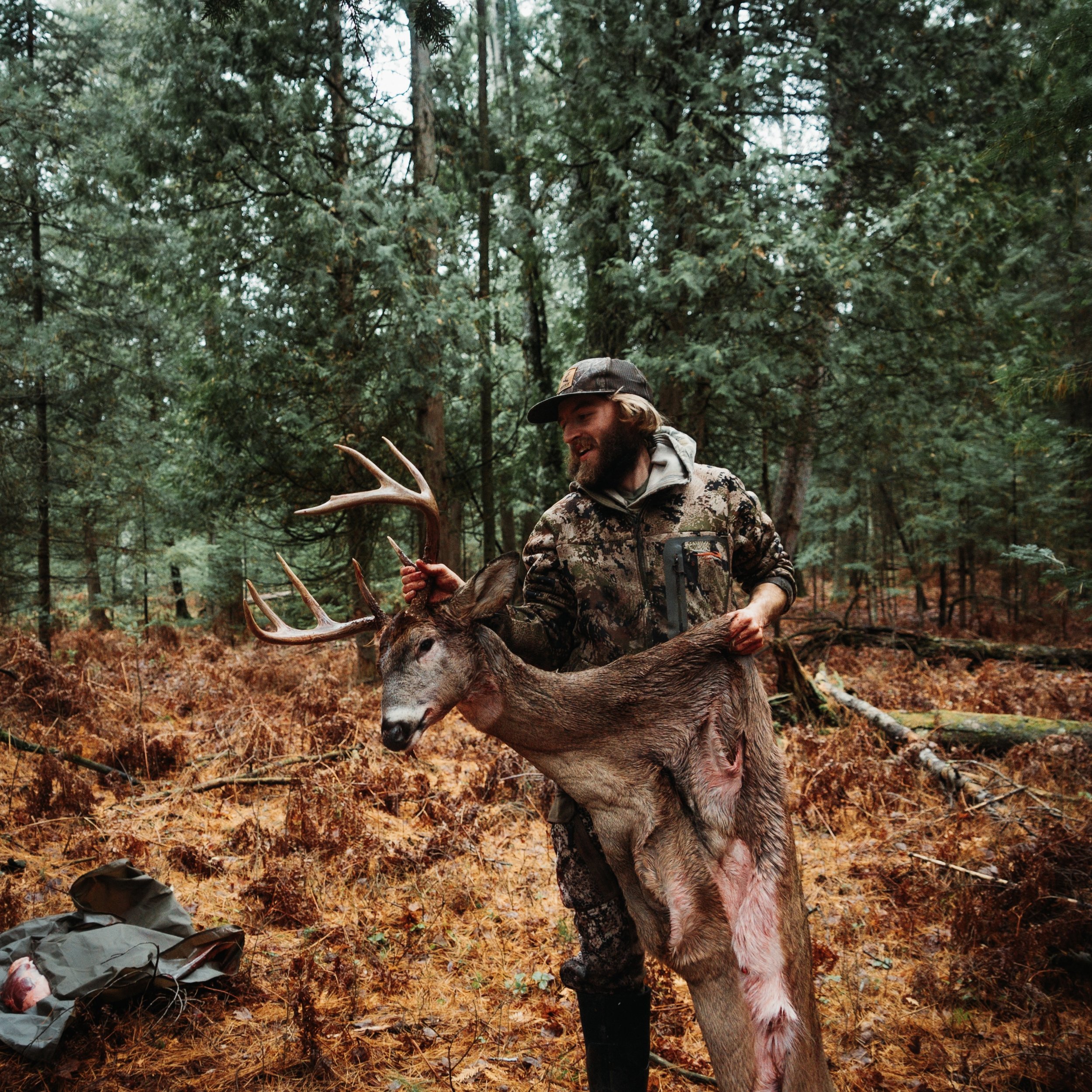 A hunter holding a large dead buck with antlers in a forested area during daytime.