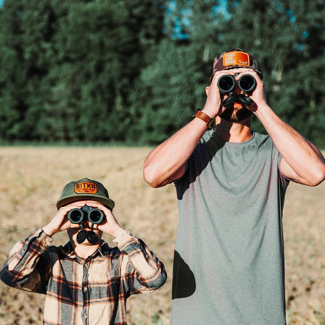 A man and a boy standing outdoors in an open field, both looking through binoculars. The man has a beard, wears a gray t-shirt, and a camouflage cap with a patch that says 'Public Land Owner.' The boy wears a plaid shirt and a green cap with a patch 
