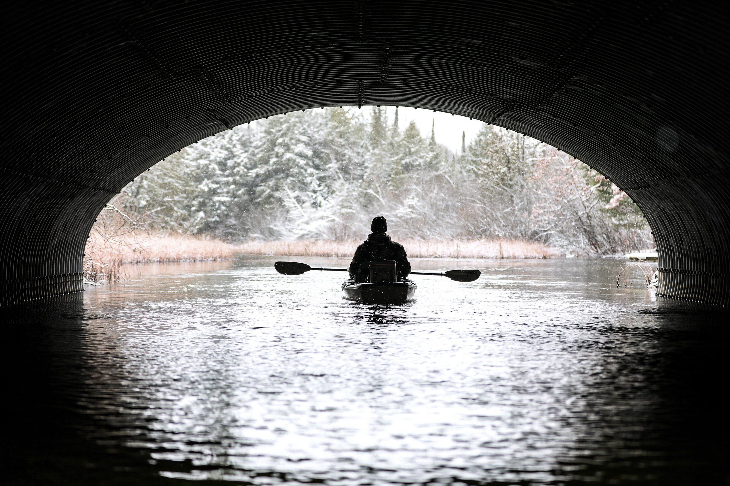 Person kayaking through a tunnel under a bridge on a river with snow-covered trees in the background.
