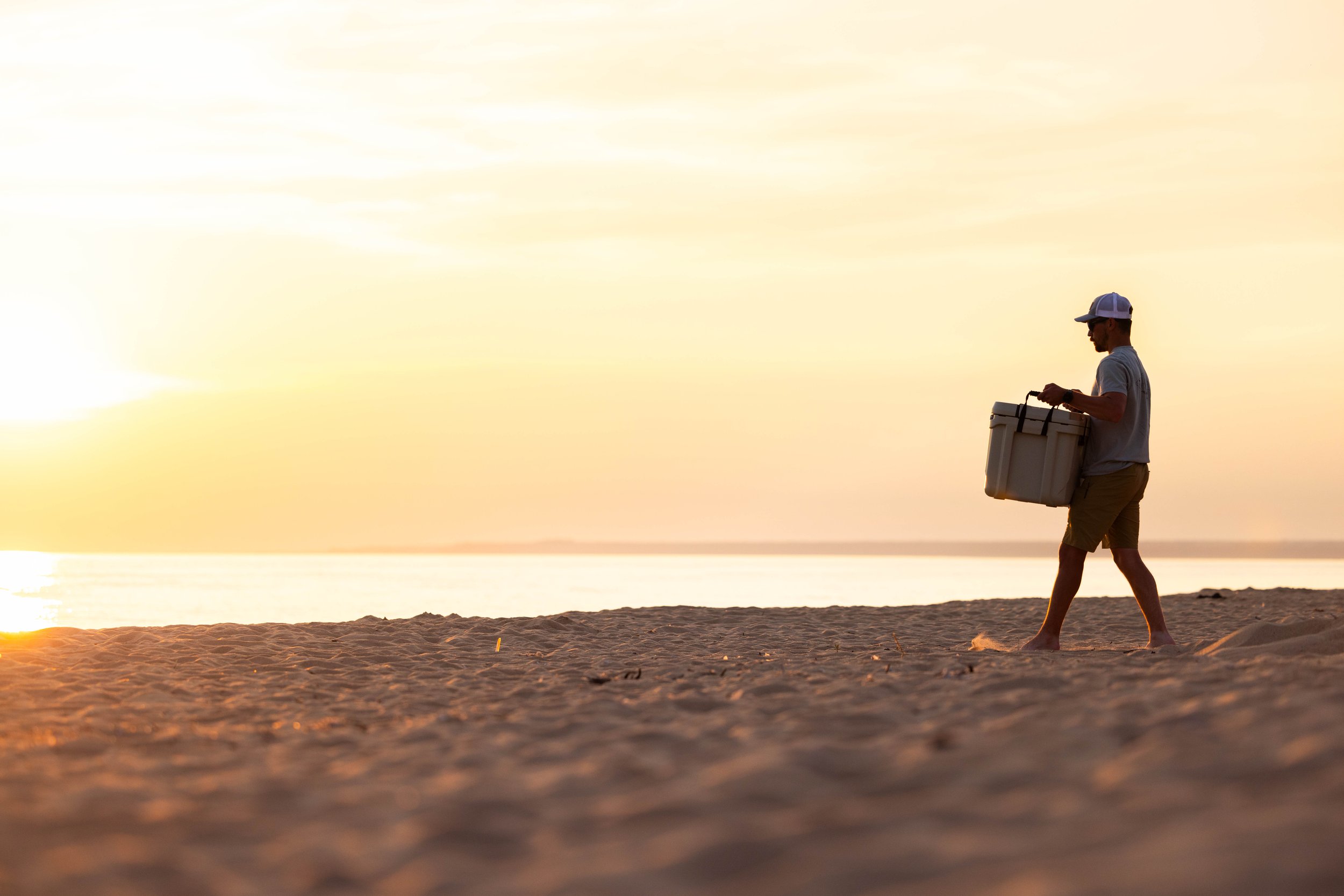 Person walking on the beach at sunset, carrying a cooler.