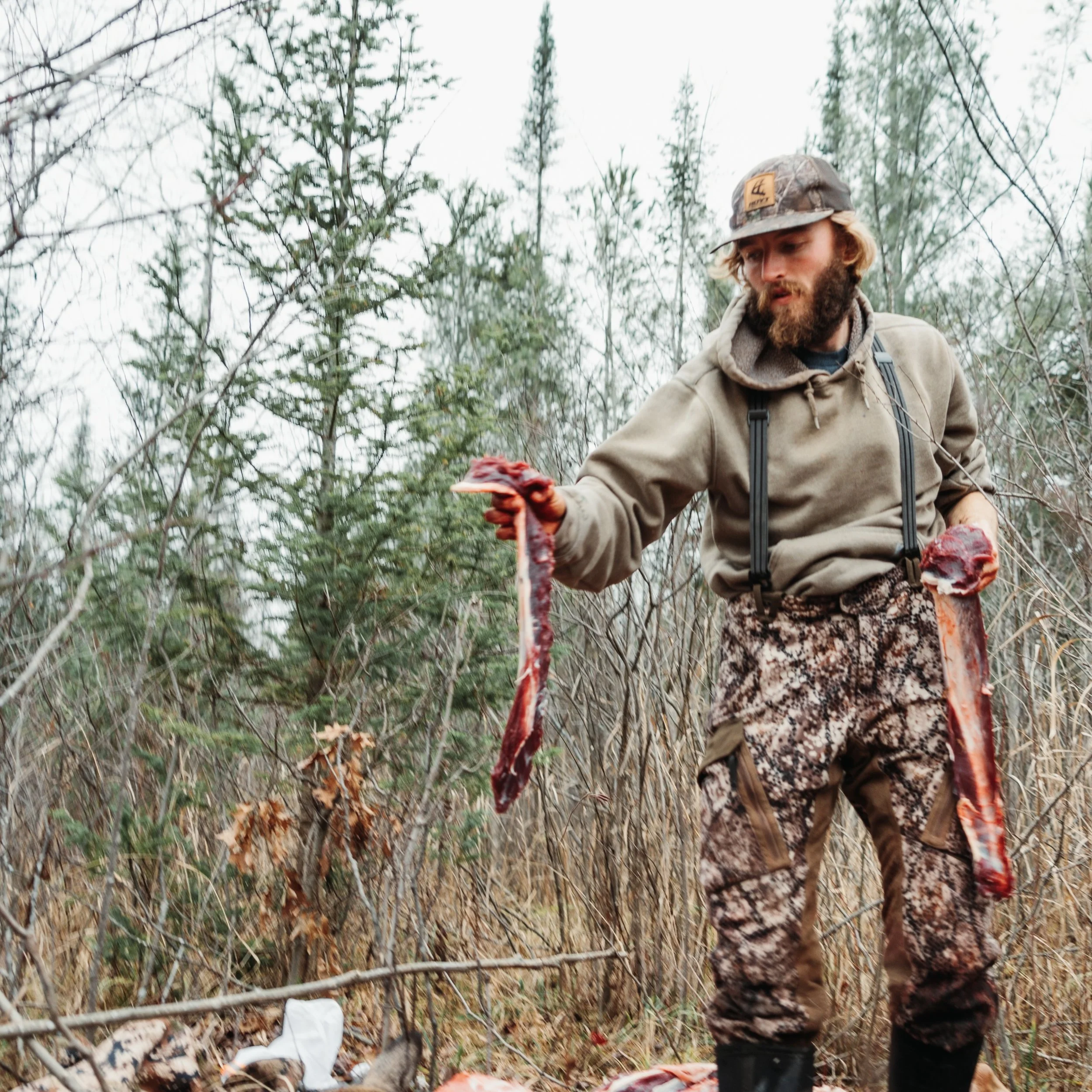 A man dressed in camouflage pants, a hoodie, and suspenders holding a freshly caught animal carcass in a wooded outdoor setting.