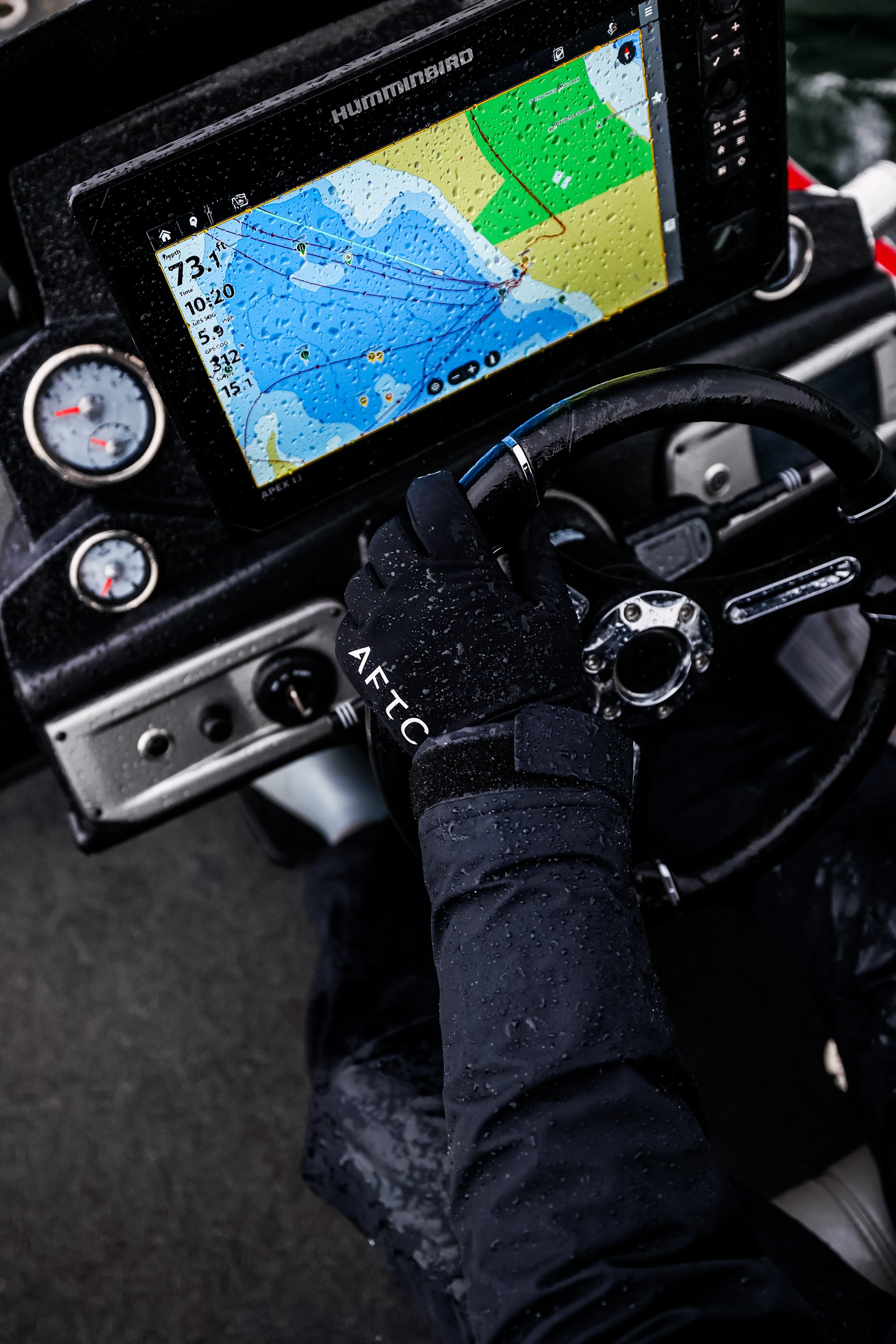 Close-up of a boat's dashboard displaying a GPS navigation screen with water depth, speed, and location markers. The water is rainy or humid, with raindrops on the screen. A person's gloved hand is on the steering wheel, and the dashboard has additio