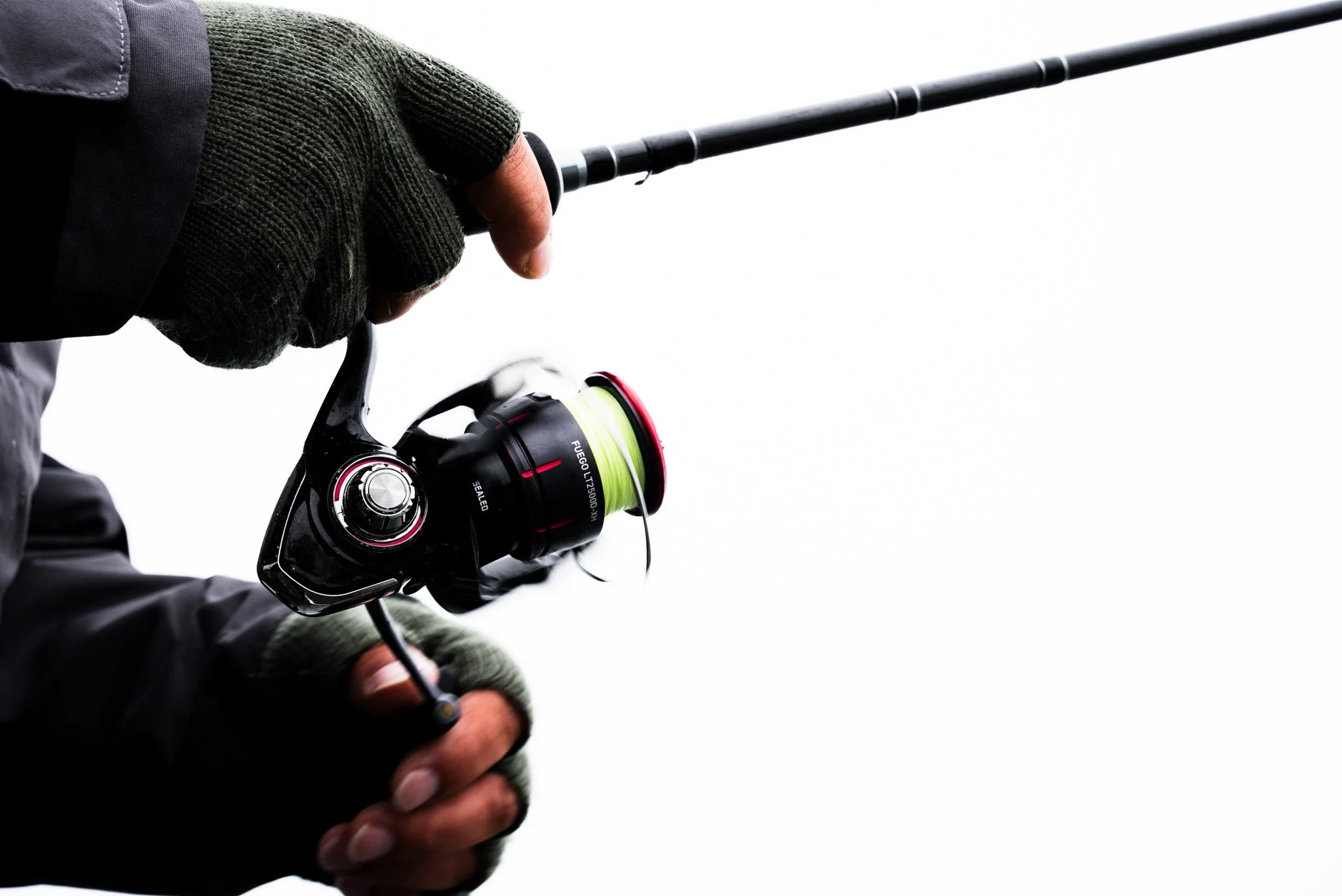 Person holding a fishing rod with a spinning reel, wearing gloves and a jacket against a plain white background.