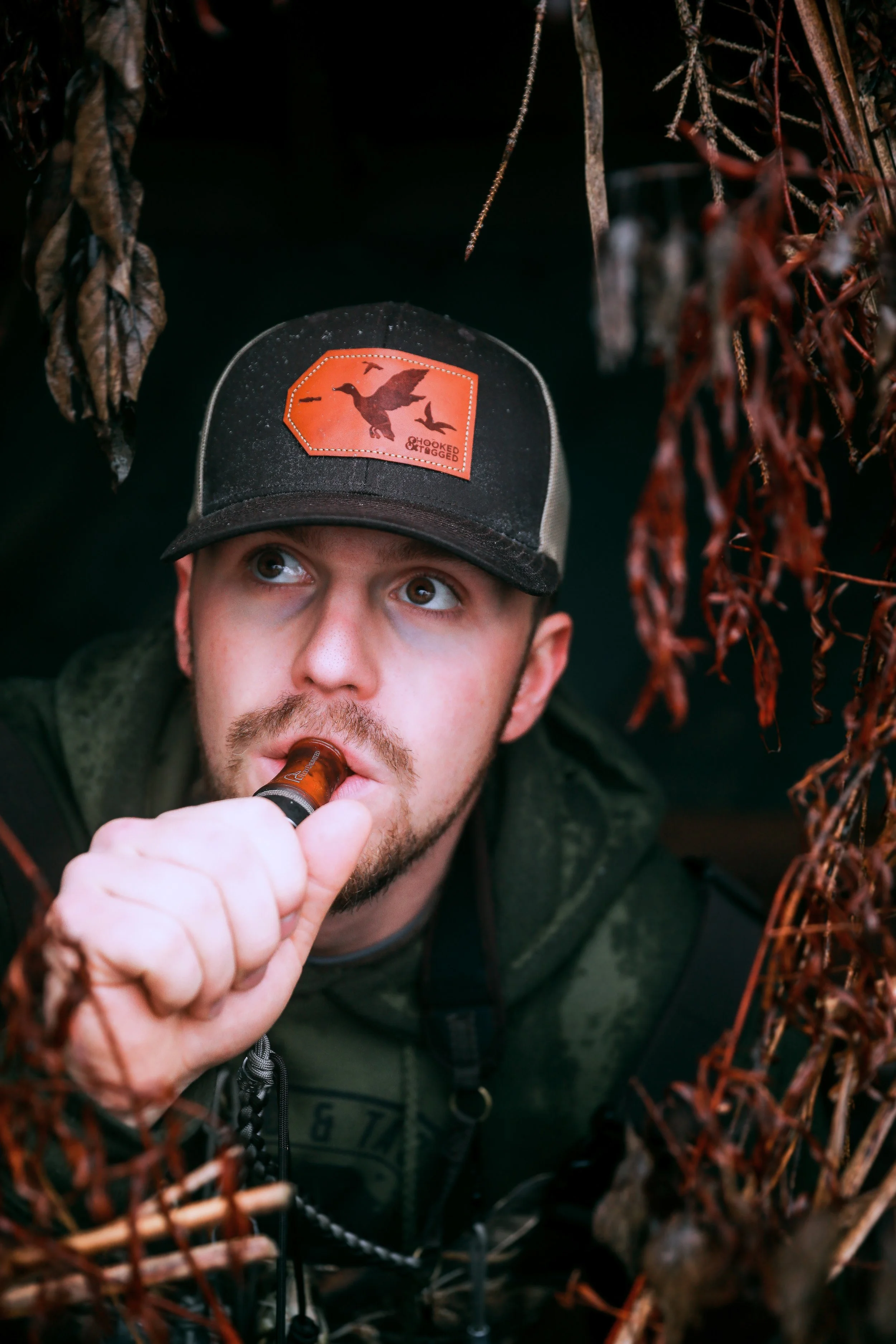A man with a beard and mustache, wearing a baseball cap and a green jacket, is holding a small bottle to his lips, possibly drinking or vaping, surrounded by dried plants or leaves.