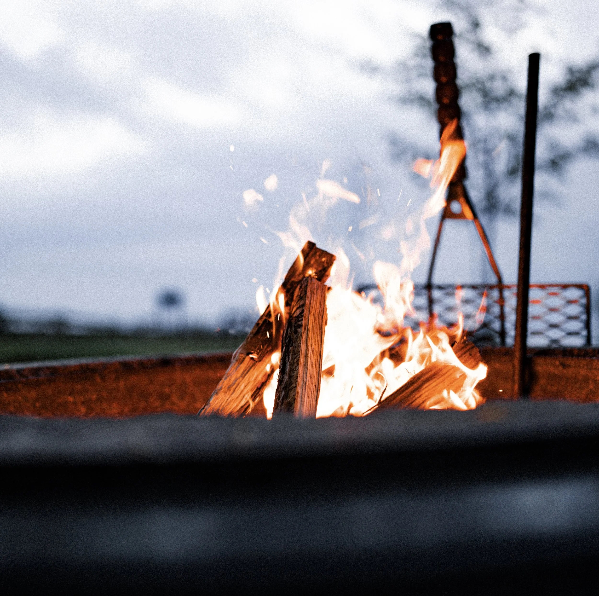 Close-up of a campfire with burning logs and orange flames, with outdoor equipment in the background.