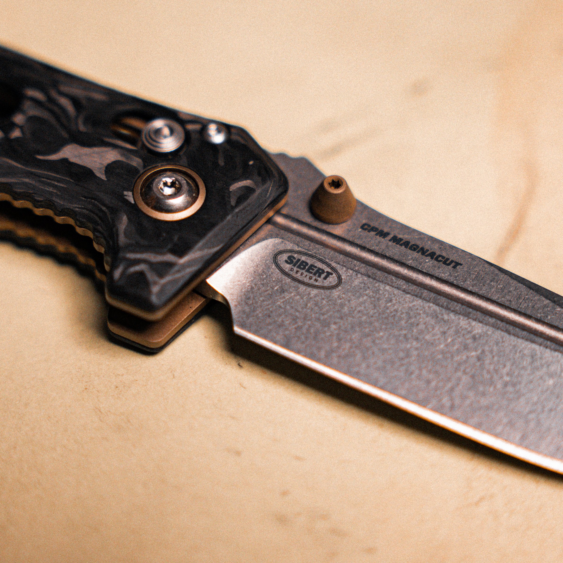 Close-up of a folding knife with a black handles and titanium blade on a beige surface.