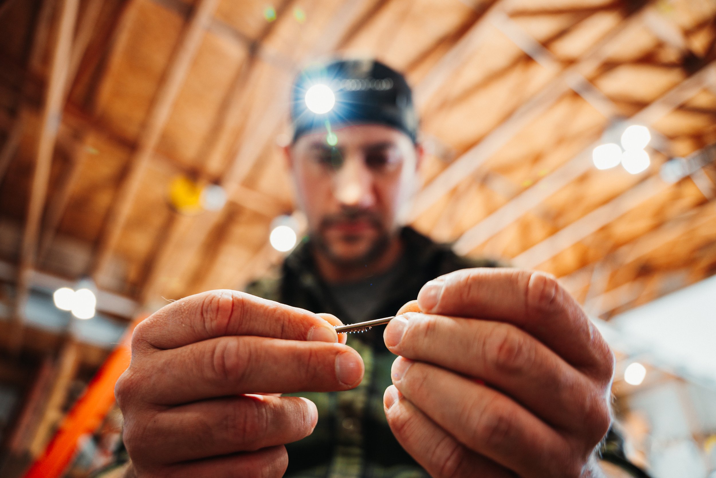 A man in a baseball cap and camouflage shirt holding a small needle or tool, with a wooden roof structure and bright lights above.