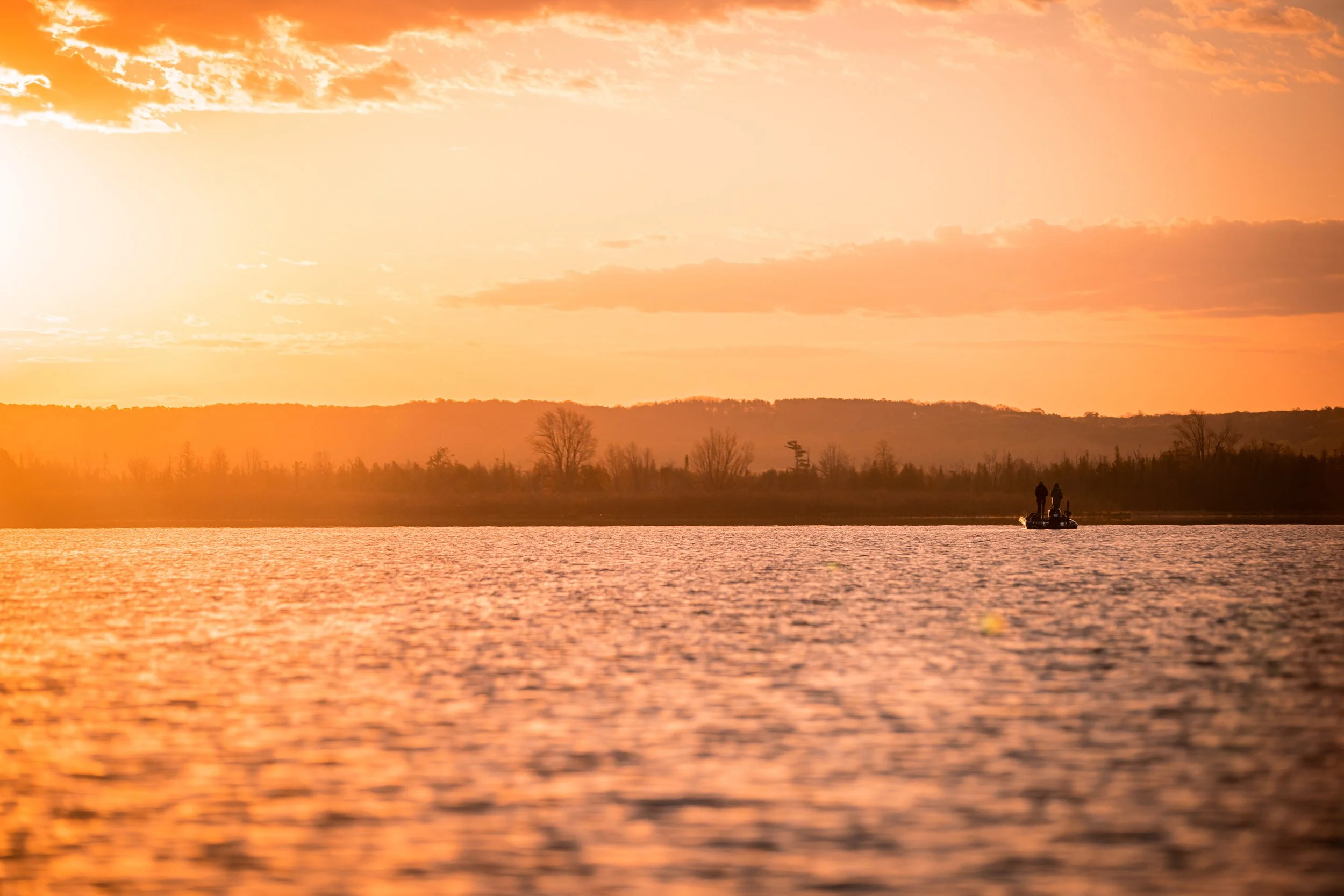 A boat with two people is on a body of water during sunset, with a distant tree-lined shore and hills in the background, under an orange and pink sky.