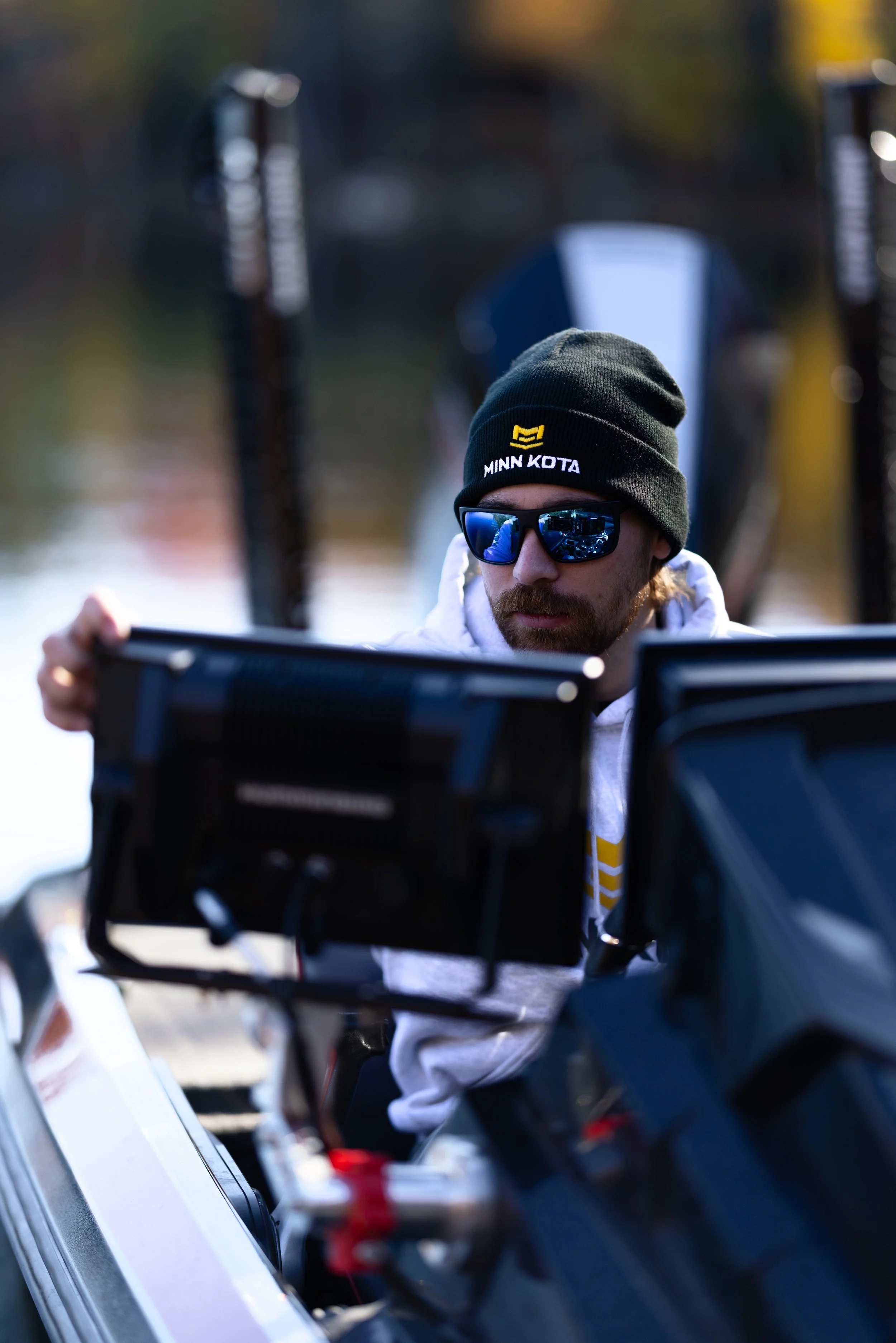 Man wearing a black beanie with "Minn Kota" logo, sunglasses, and a white hoodie, focusing on a boat control panel outdoors.