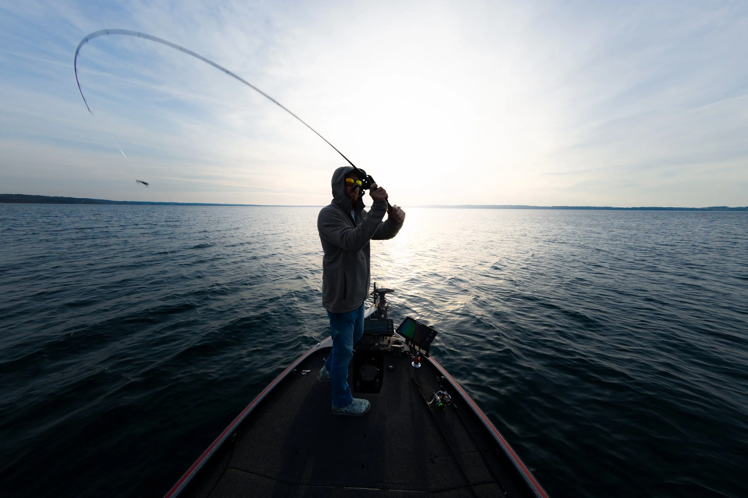 Person fishing on a boat in a large body of water during sunset or sunrise.