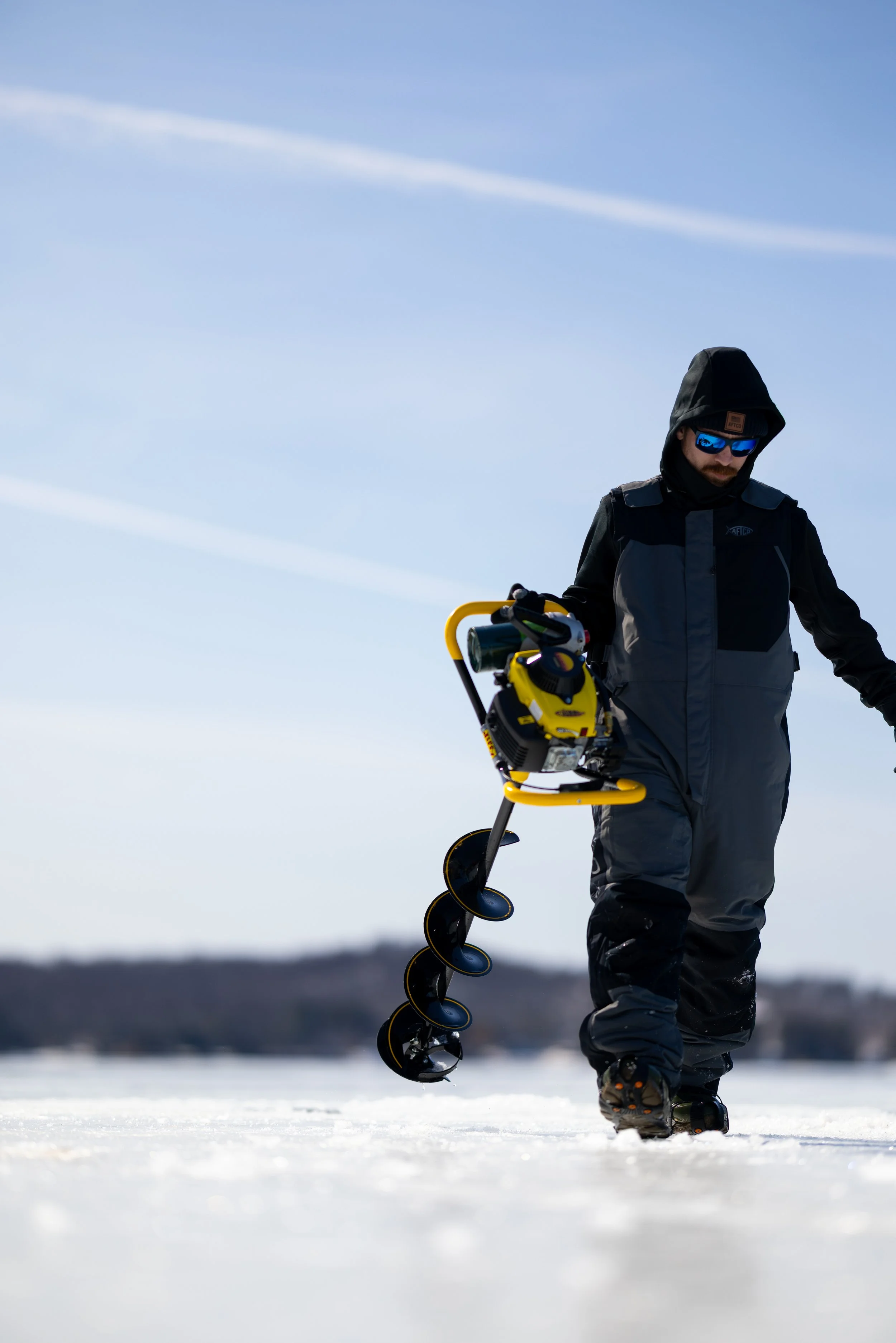 Man dressed in winter clothing walking on snow while holding a yellow ice auger.