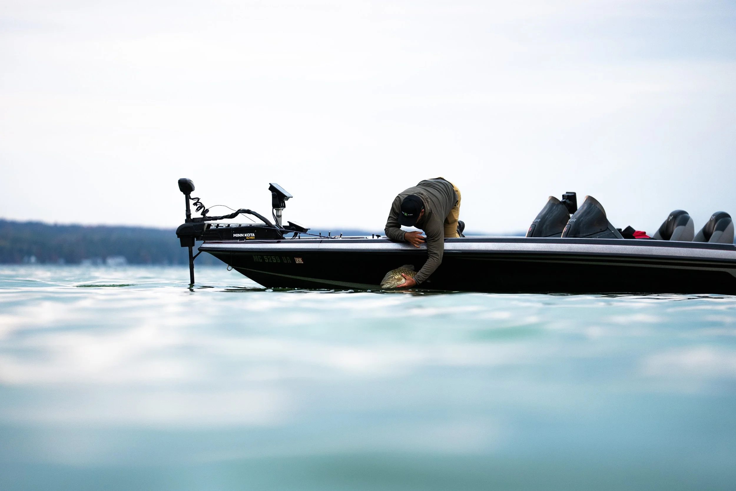 A man leaning over the side of a black fishing boat holding a fish in his hand on a calm lake with a faint shoreline in the background.