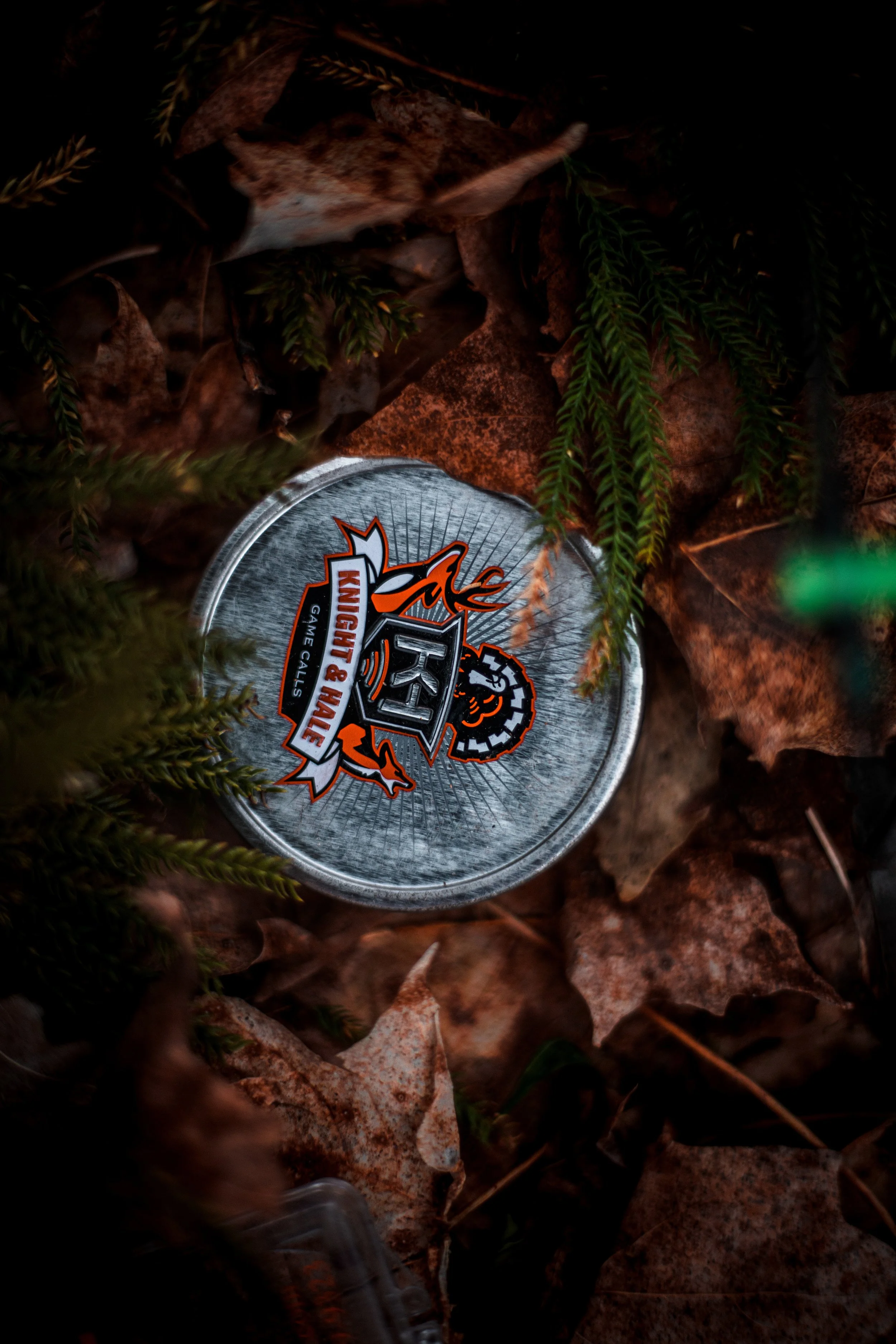 An empty metal can with a hunting and game call logo is lying on the ground amidst brown fallen leaves and green pine branches.