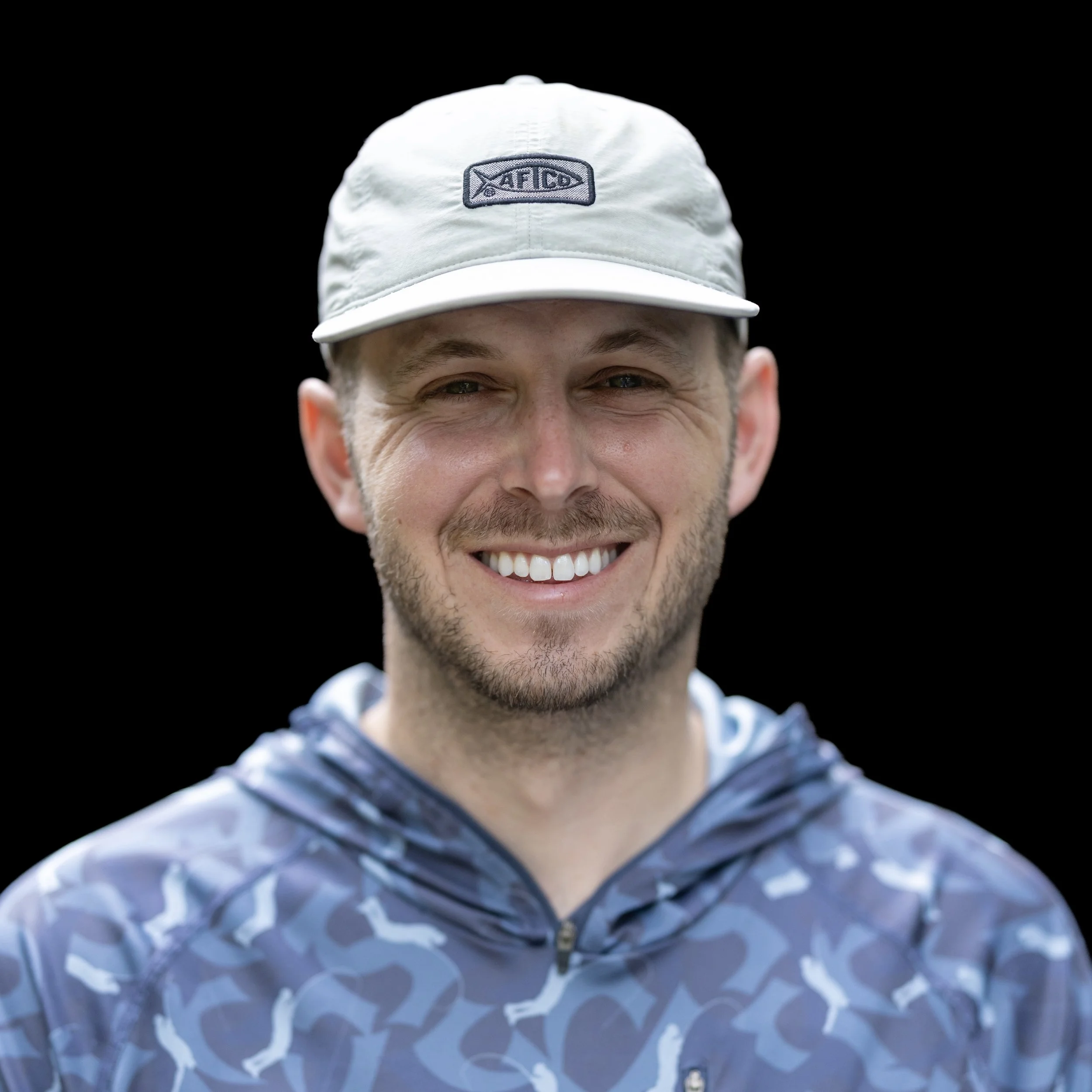 Close-up of a smiling man with a beard wearing a light-colored hat and a patterned jacket against a black background.