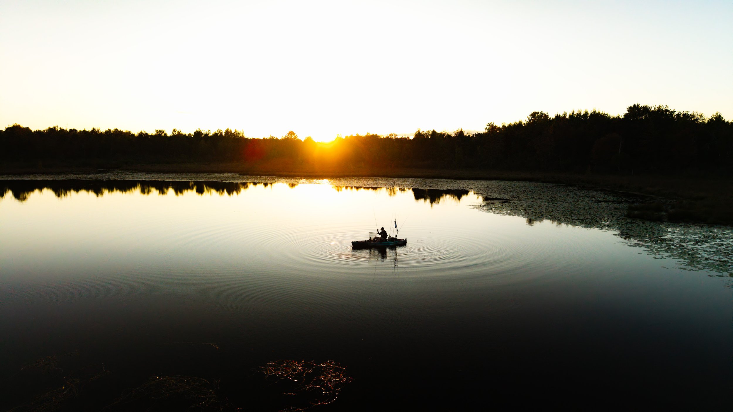 A person fishing from a small boat on a calm lake at sunset, with trees silhouetted in the background.