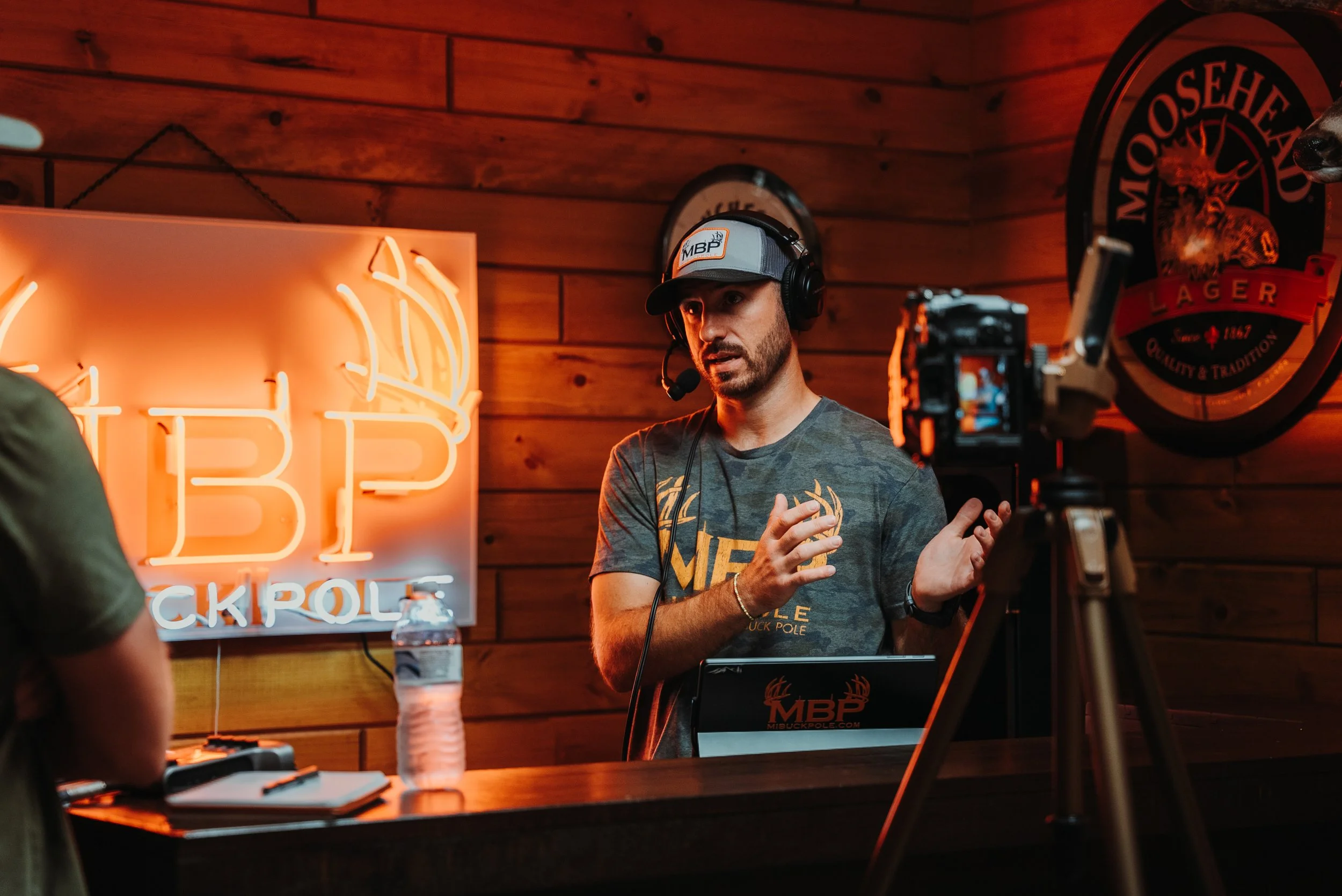 A man wearing a gray MBP cap, headset, and camouflage T-shirt speaking into a microphone at a wooden bar with a neon sign and Moosehead Lager logo in the background.