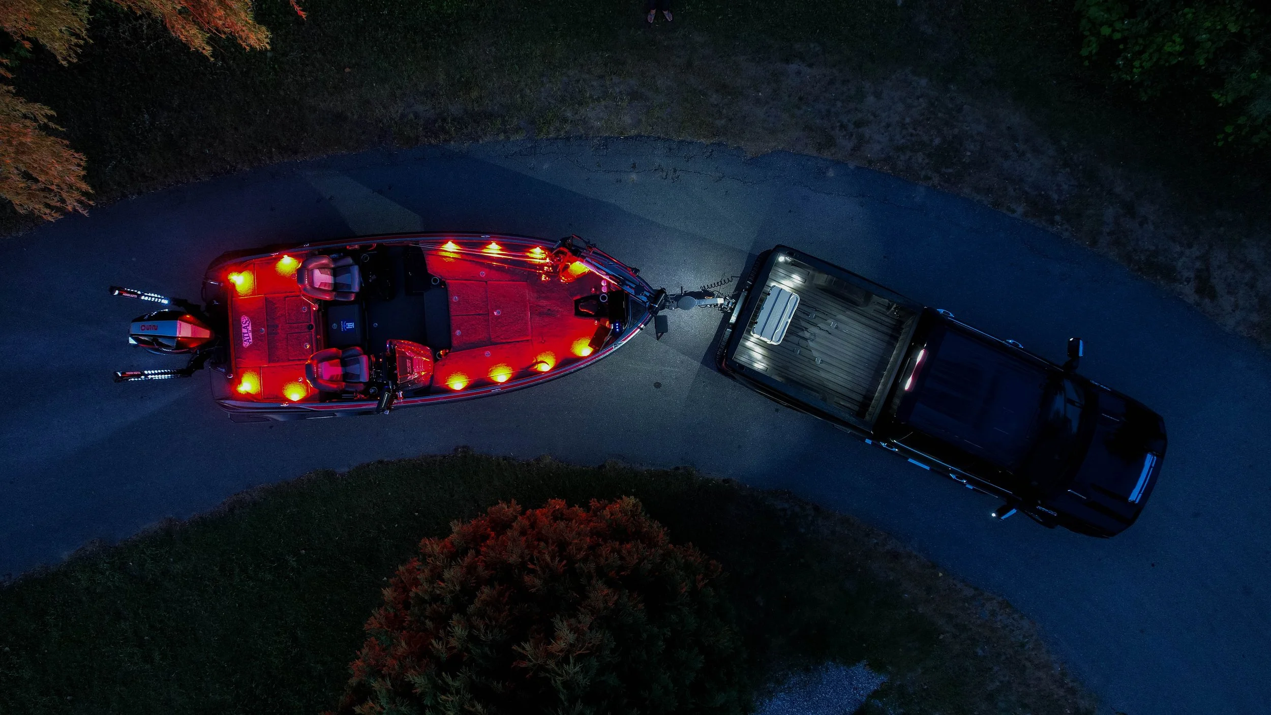 An aerial view of a boat attached to a pickup truck on a dark road at night, with the boat's onboard lights illuminating.