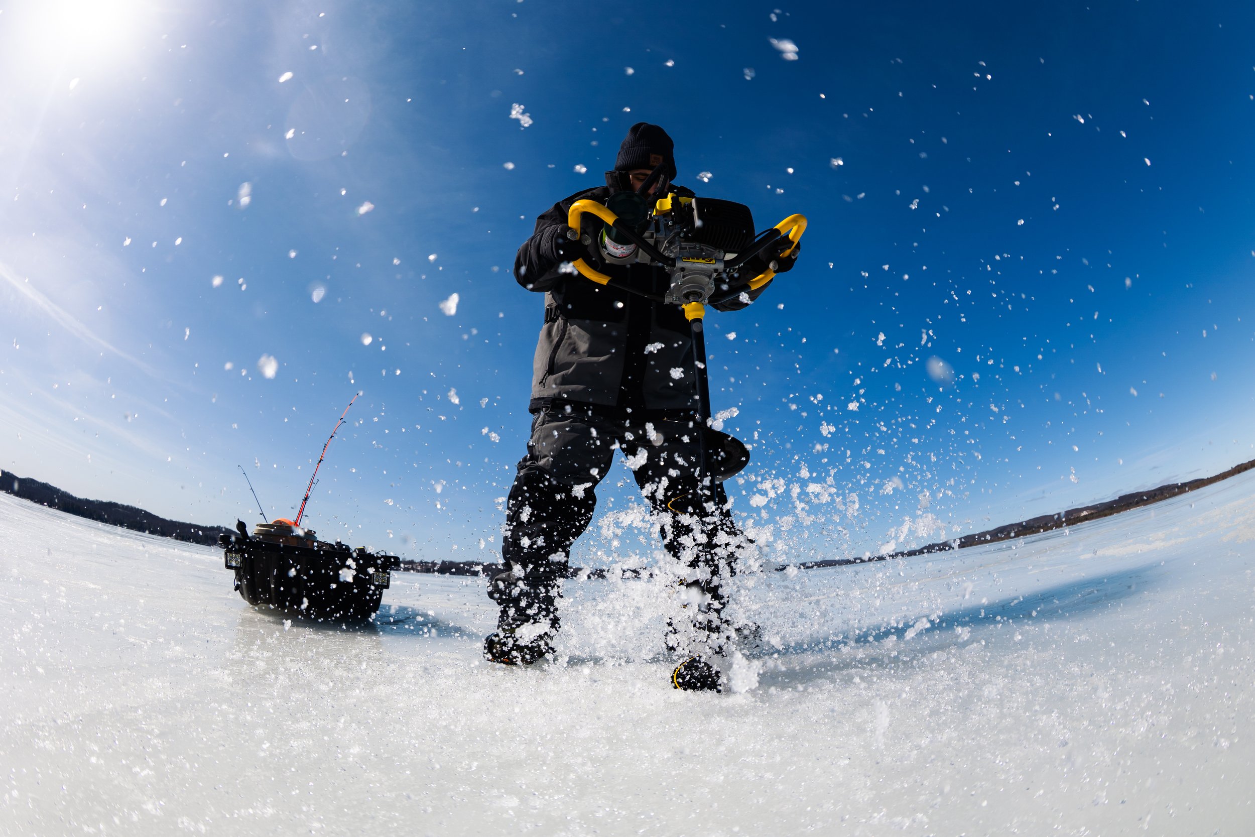 Person operating ice fishing equipment on a frozen lake, with snow and ice flying in the air, under a clear blue sky.