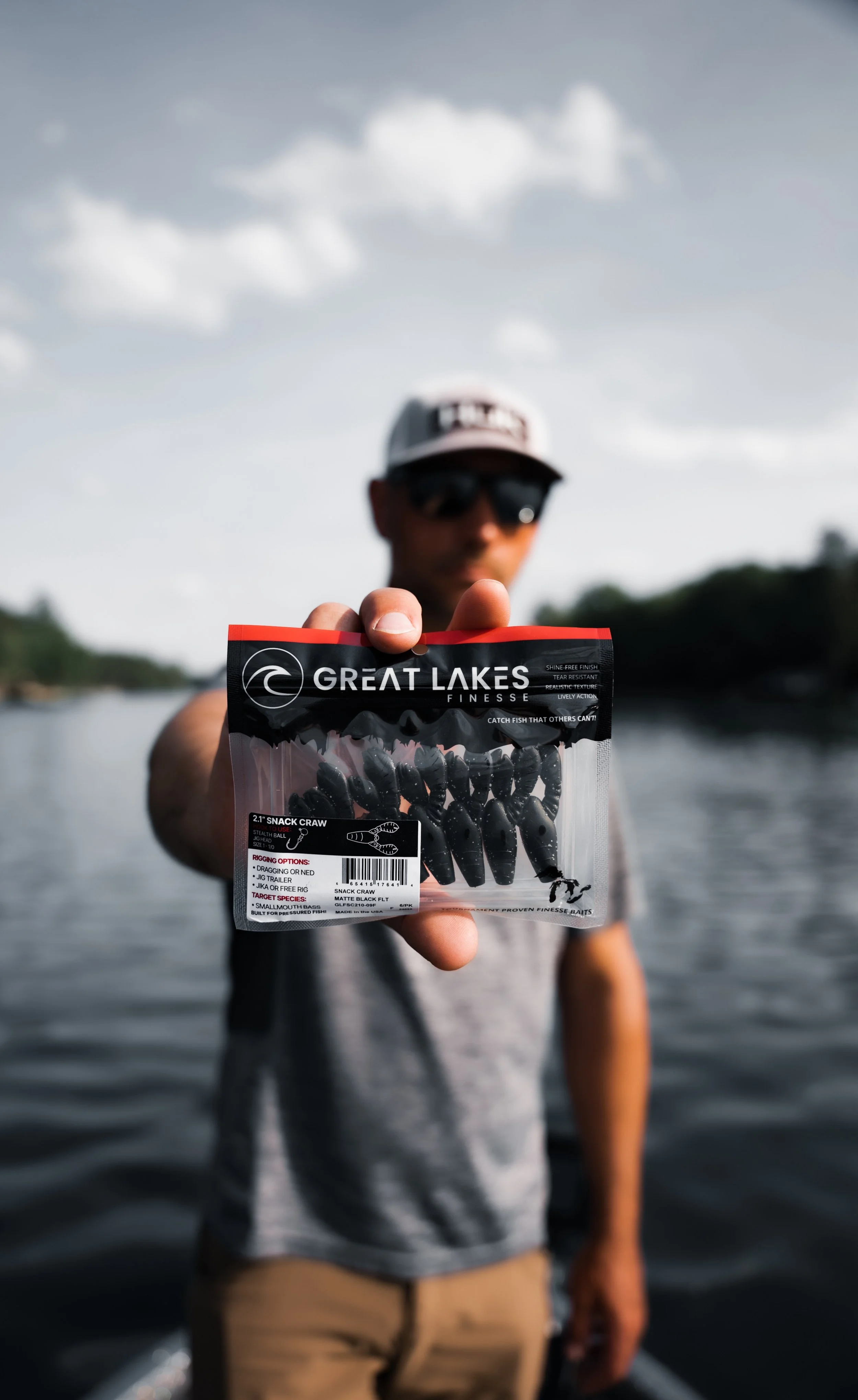 A man standing near a body of water, holding a package of fishing soft bait lures labeled "Great Lakes Finesse Snack Craw" with a blurred background of trees and cloudy sky.