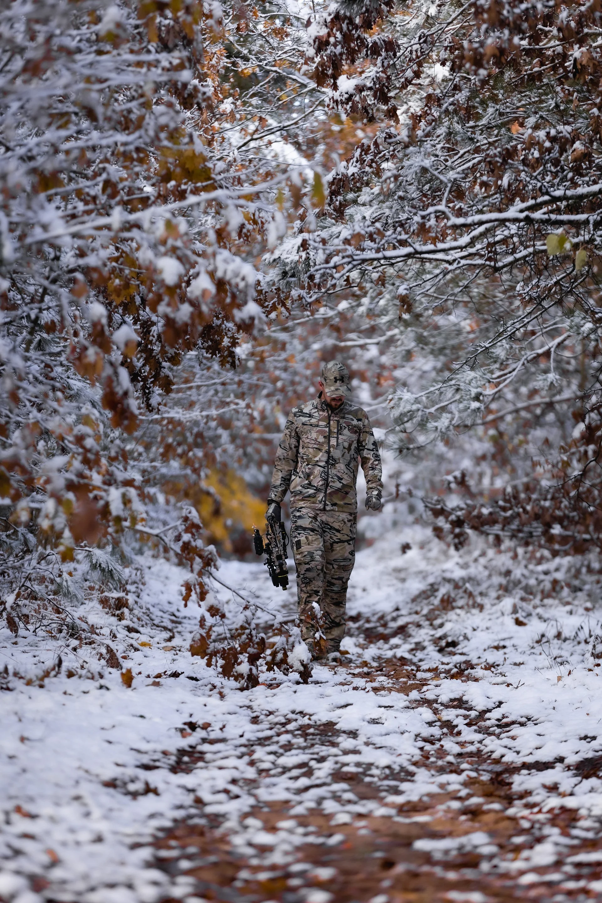 A person wearing camouflage military clothing walking through a snow-covered forest path, holding binoculars.