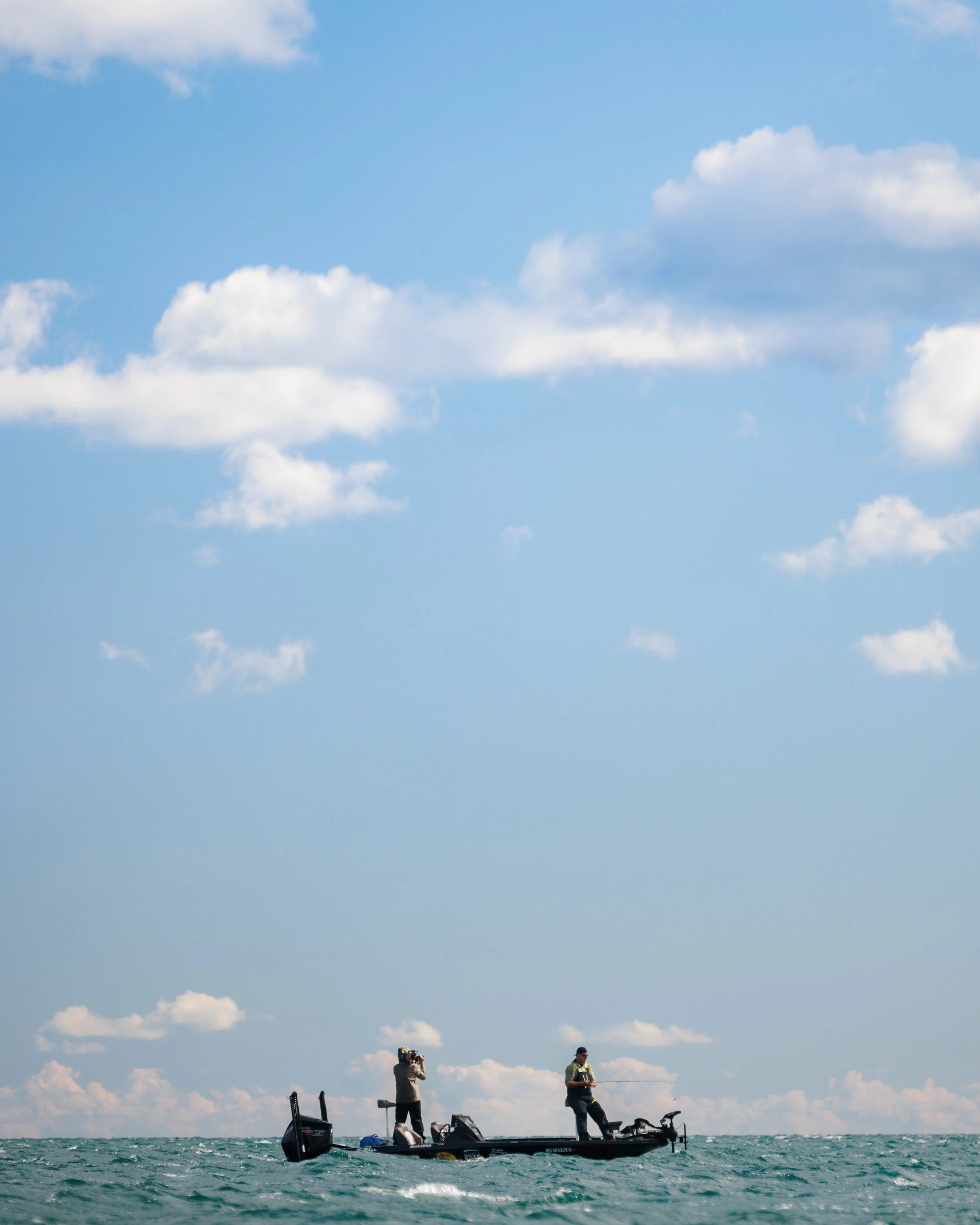 Two men fishing on a small boat in the ocean under a blue sky with scattered clouds.