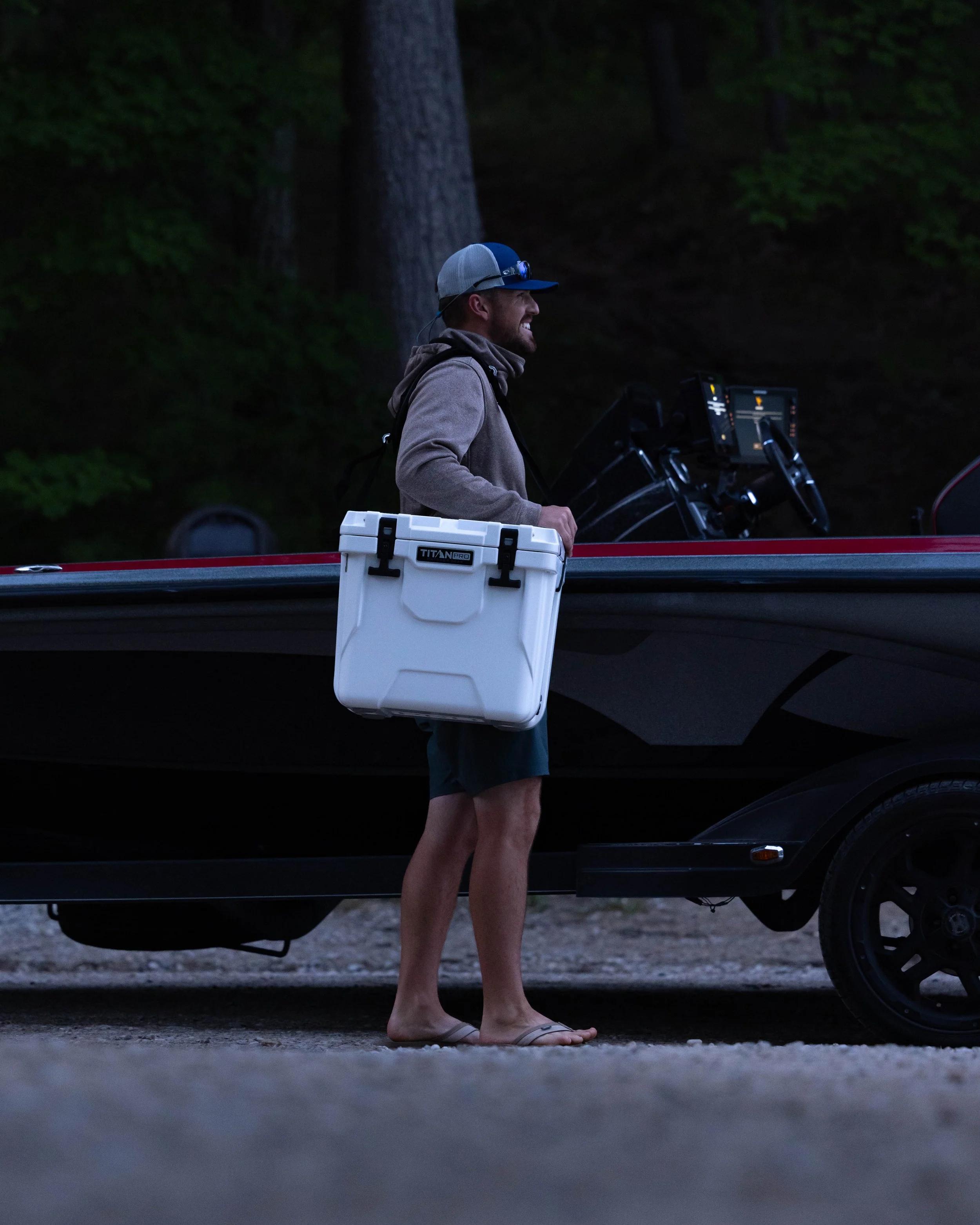 A man standing next to a boat at dusk, smiling, wearing a baseball cap, hoodie, and flip-flops, with a cooler in one hand and a boat control panel in front of him.