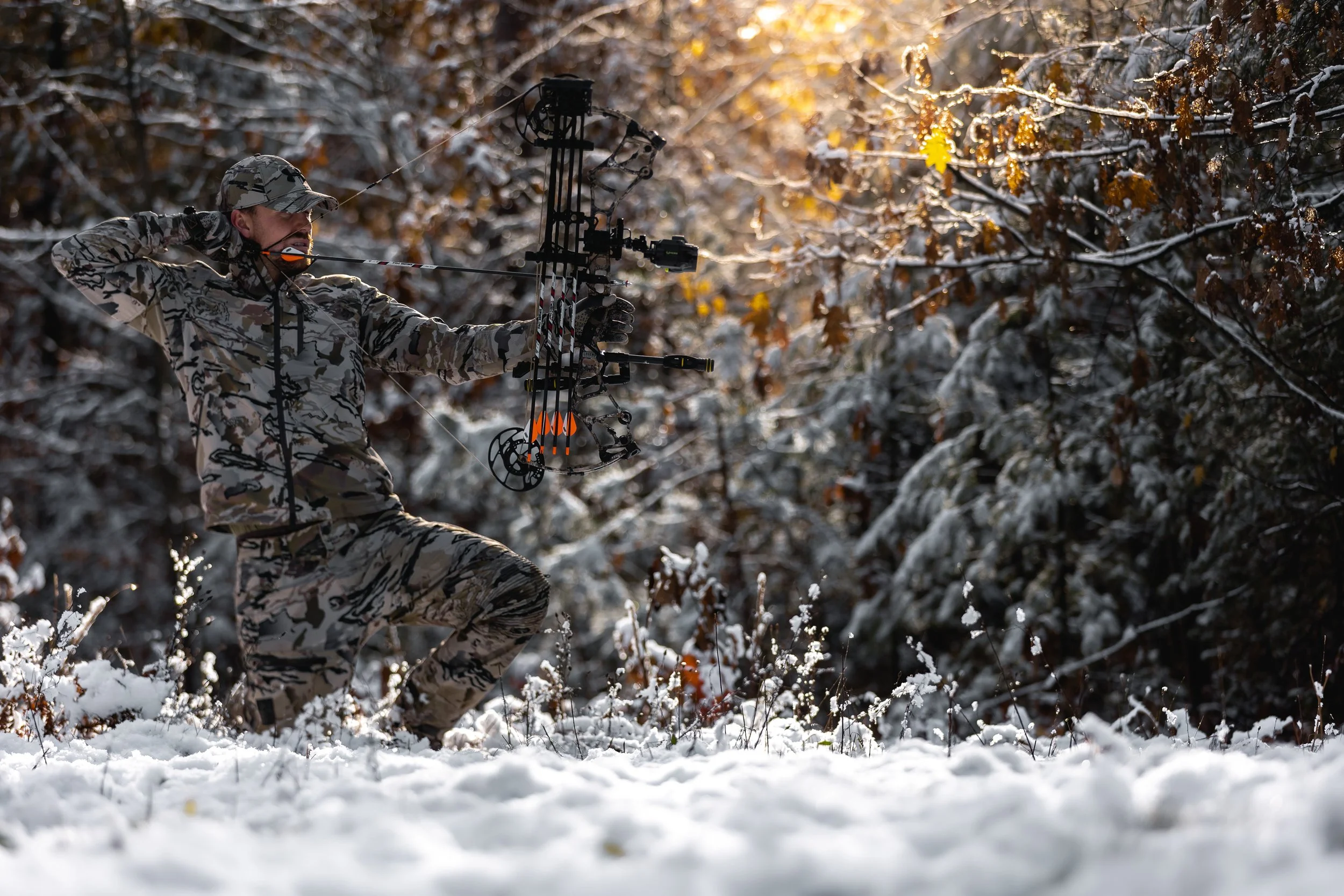 A man in camouflage clothing aiming a bow and arrow in a snowy forest during daylight.