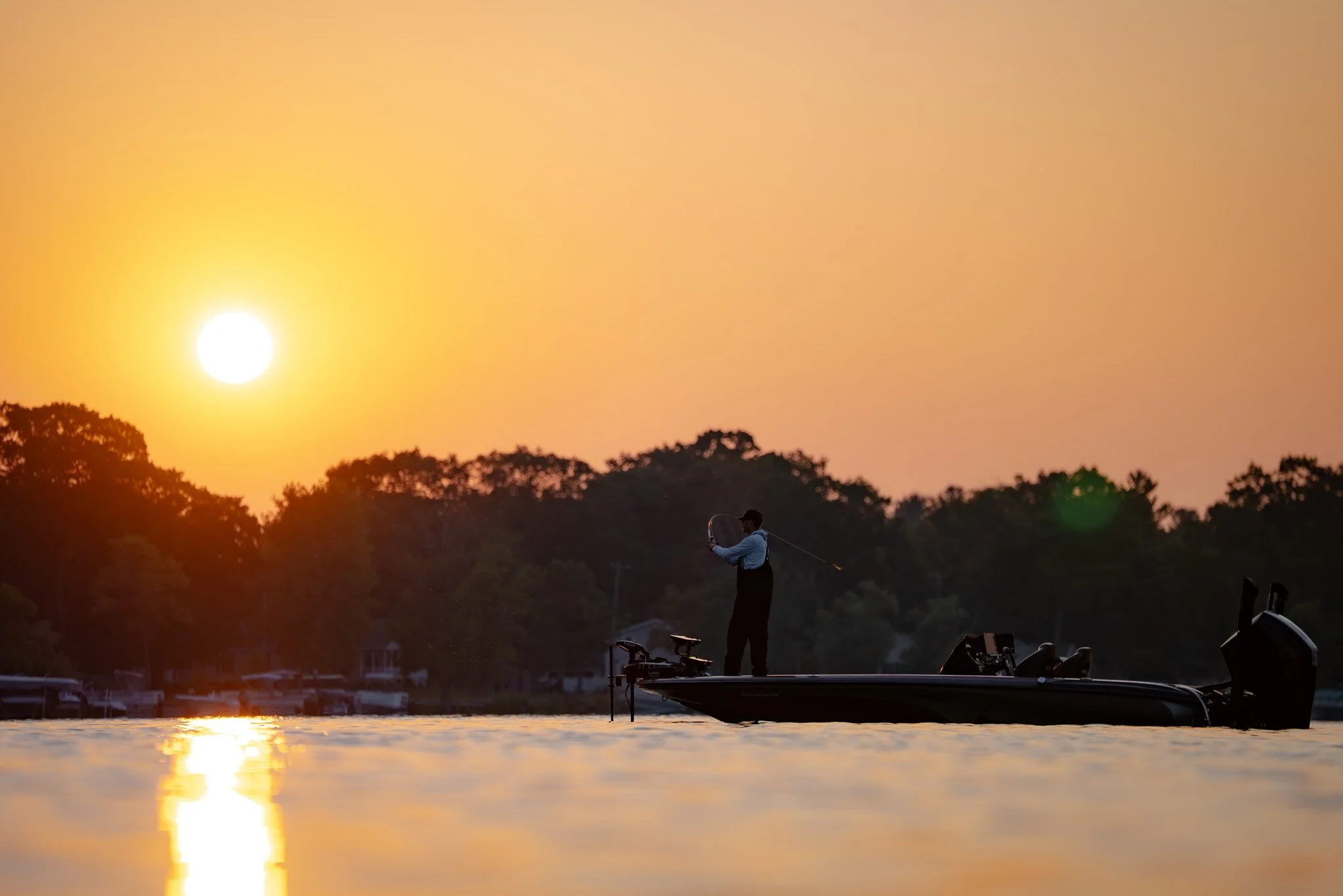 A person fishing on a boat during sunset on a calm river with a shoreline lined with trees in the background.