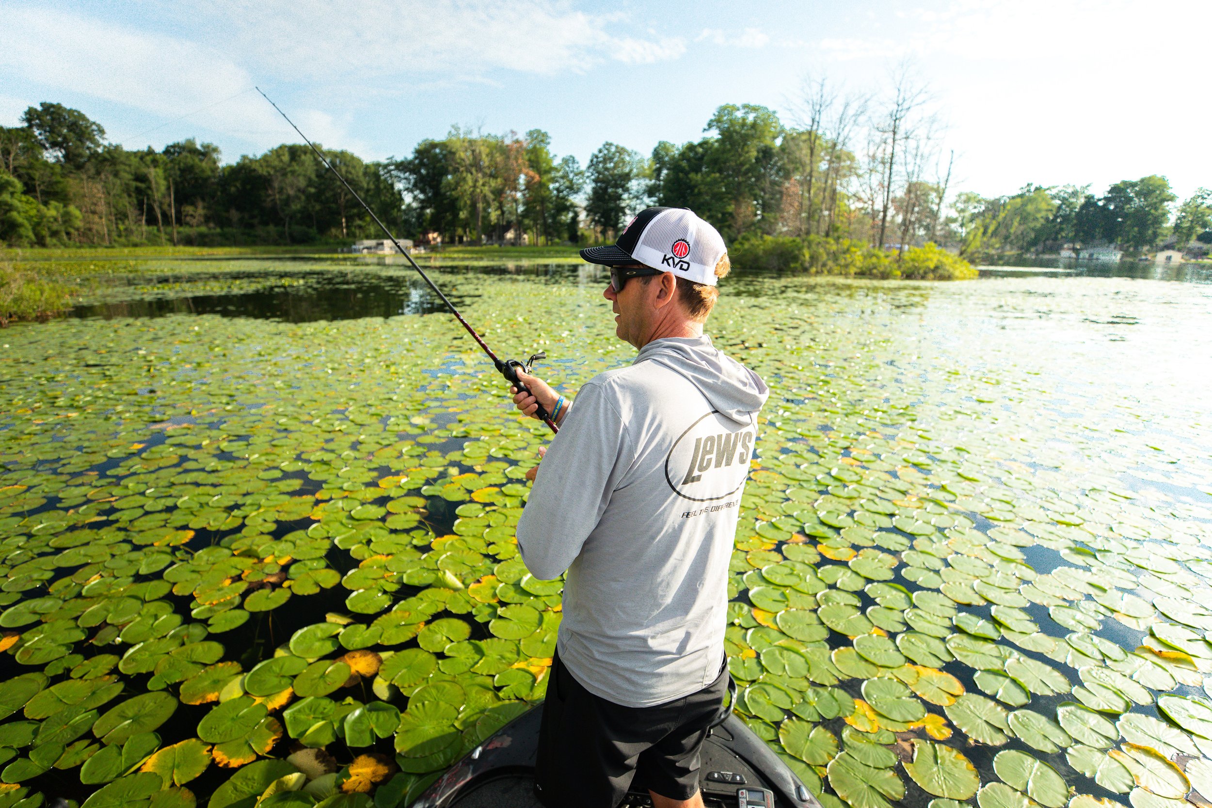 A man in a gray hoodie and a white cap fishing in a pond filled with lily pads.