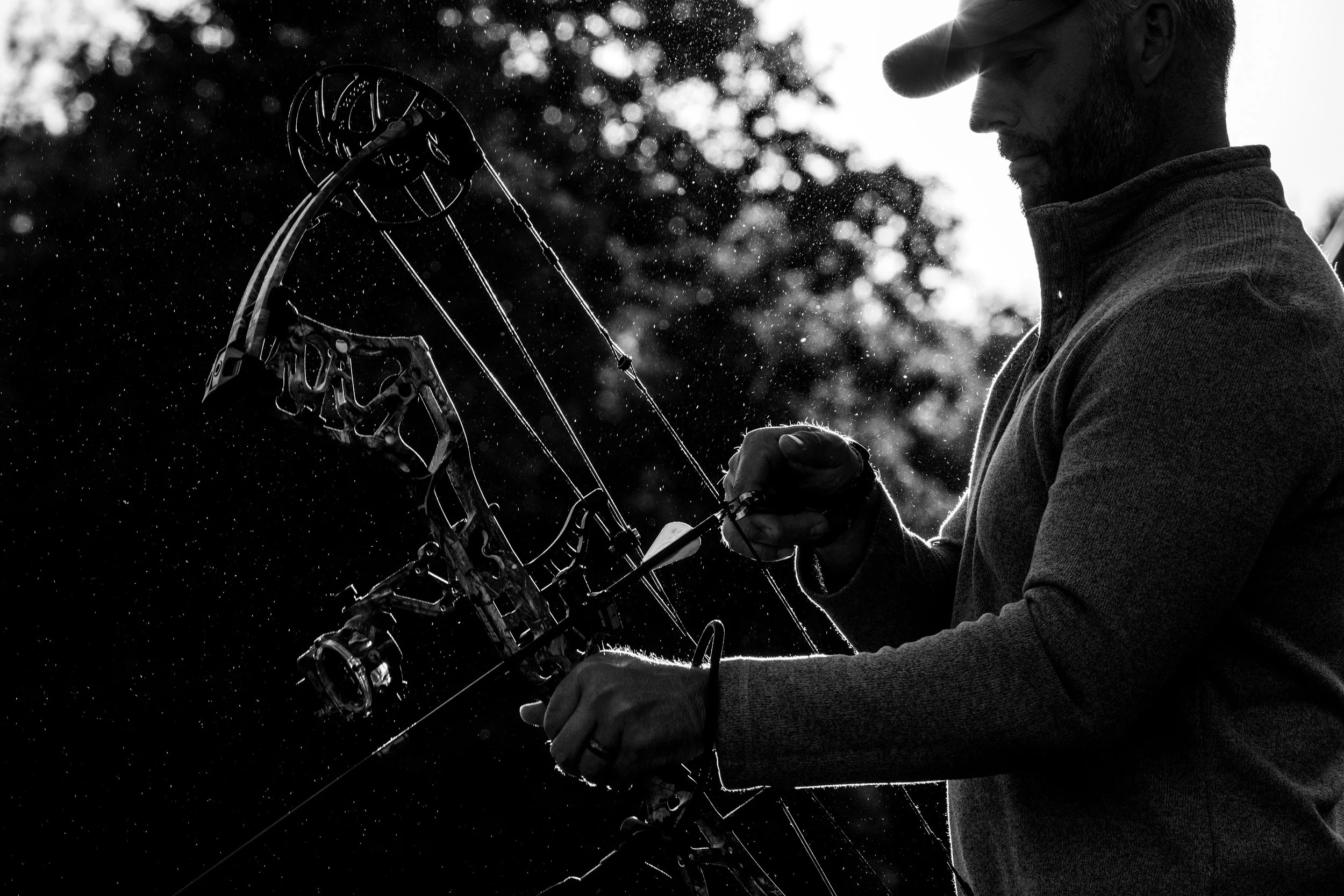 A man holding a compound bow with water droplets flying in the air, backlit by the sunlight.