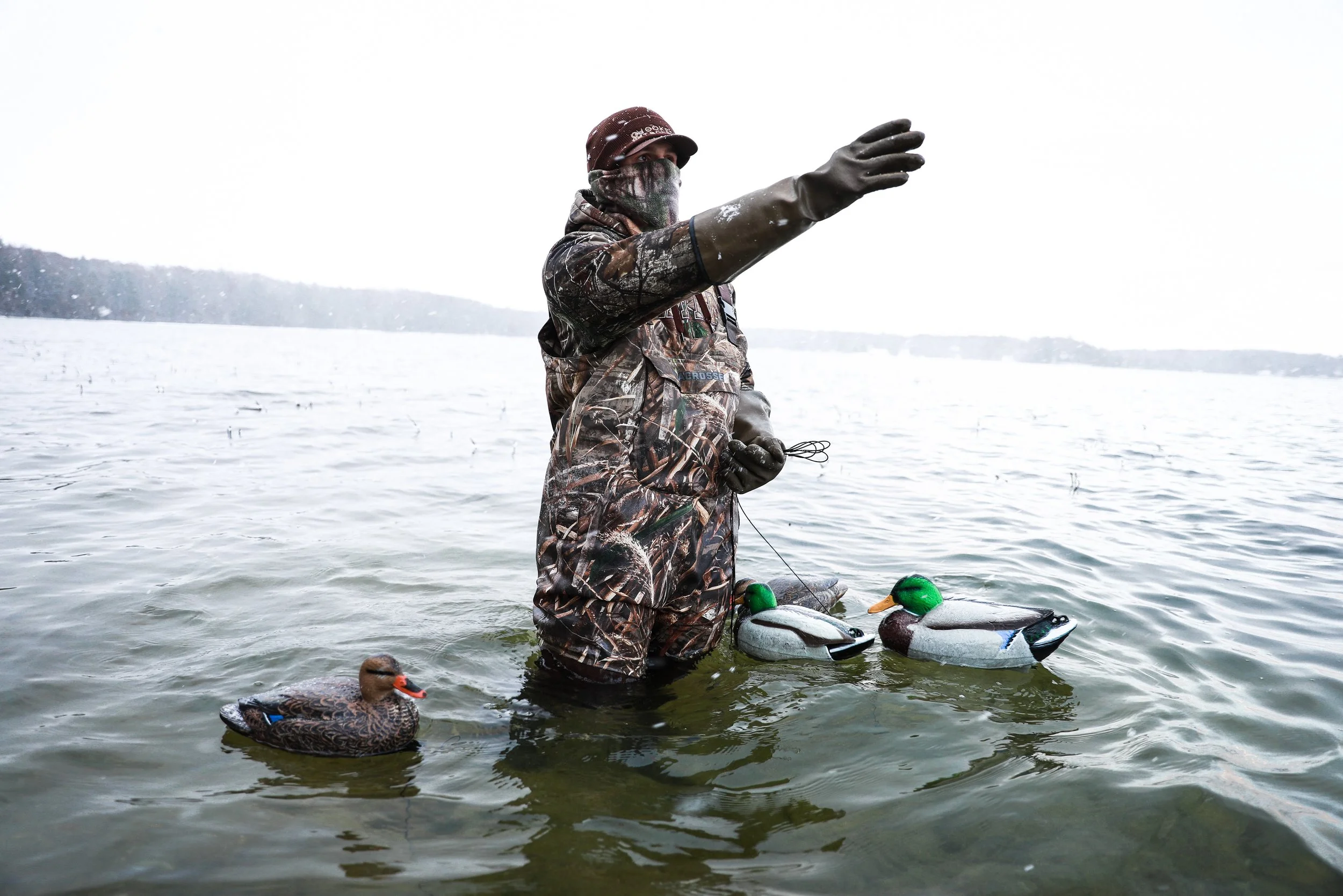 Person in camouflage clothing, wearing gloves and a face covering, standing in water holding a pair of floating decoys resembling ducks, with one real duck floating nearby, on a lake with a distant shoreline under an overcast sky.