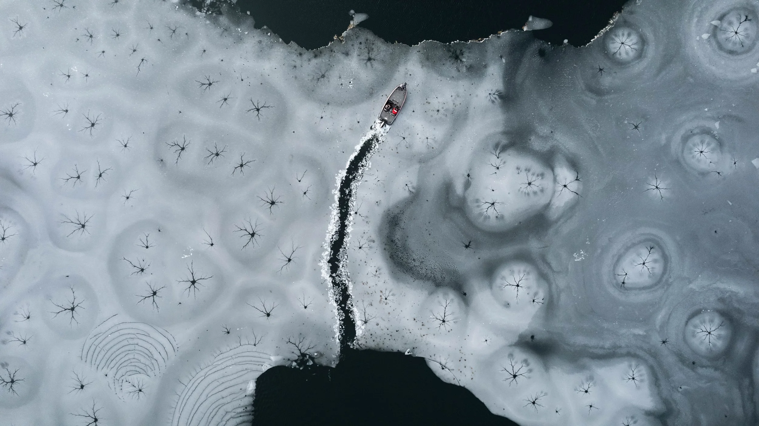 An aerial view of a boat on icy water surrounded by cracked ice with snowflakes or ice formations on the surface.