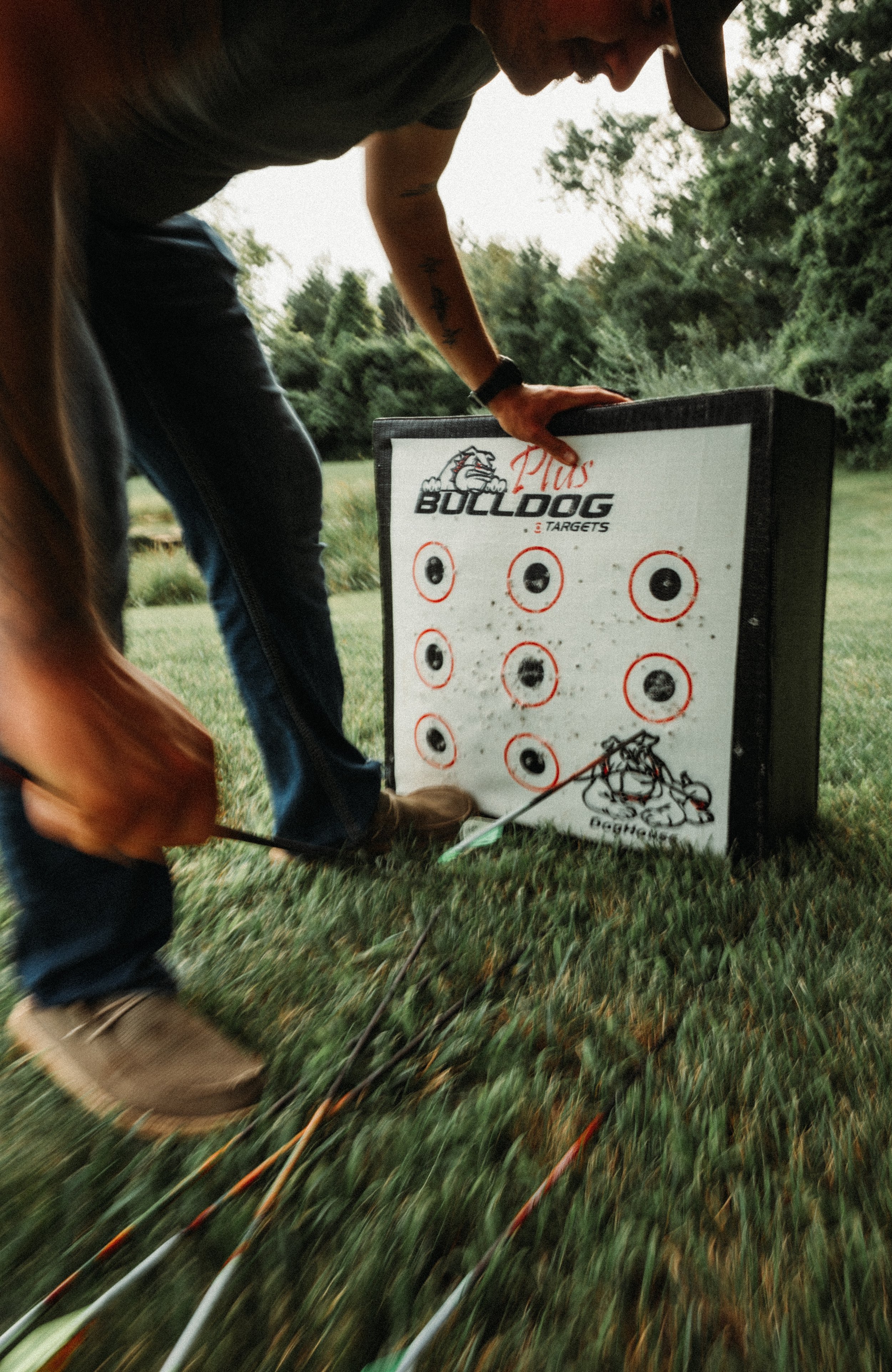 A person in a gray shirt and baseball cap leaning over a target board outdoors, holding a bow, with multiple arrows embedded in the targets.