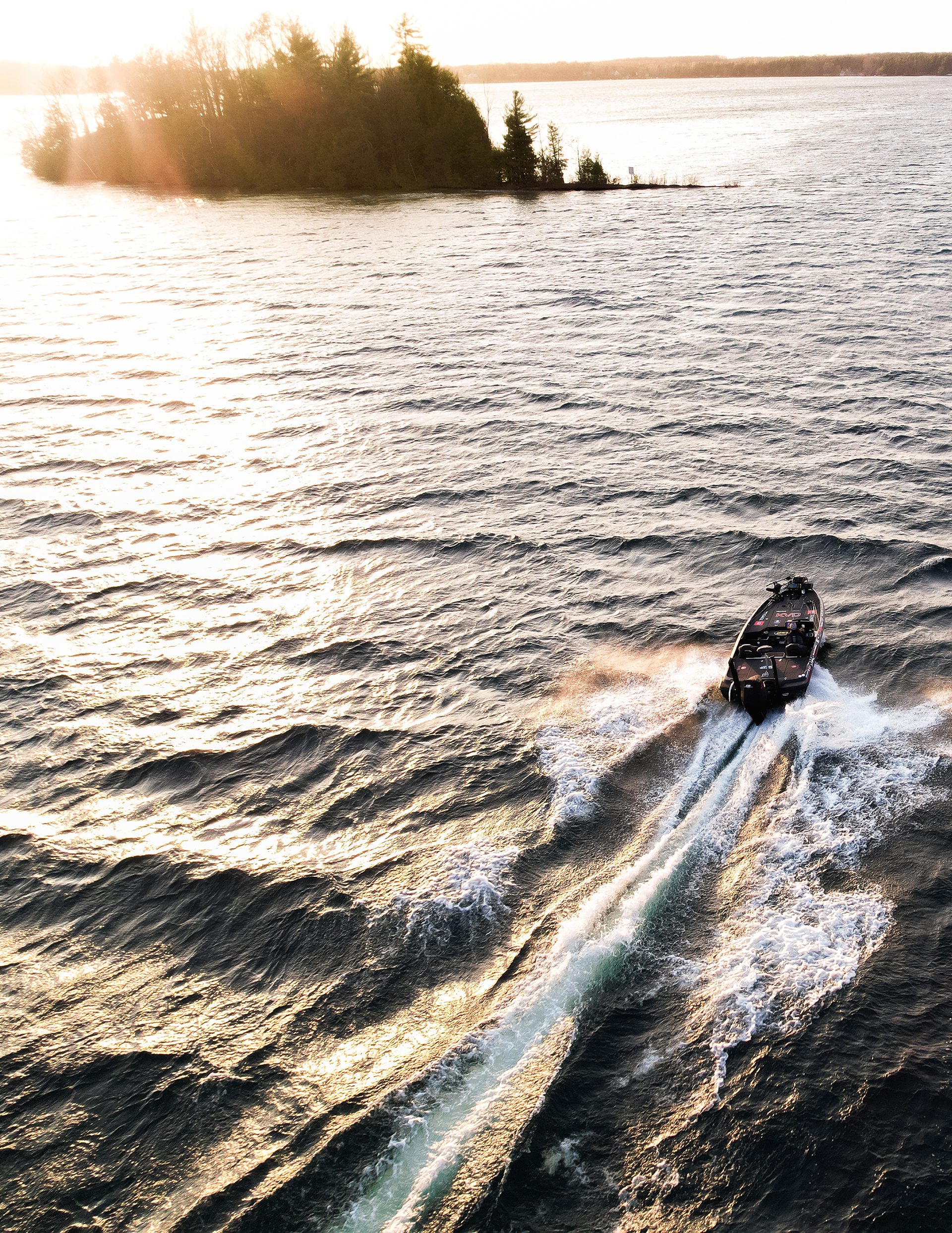 A motorboat speeding across a large body of water at sunset, with a small island and trees in the background.