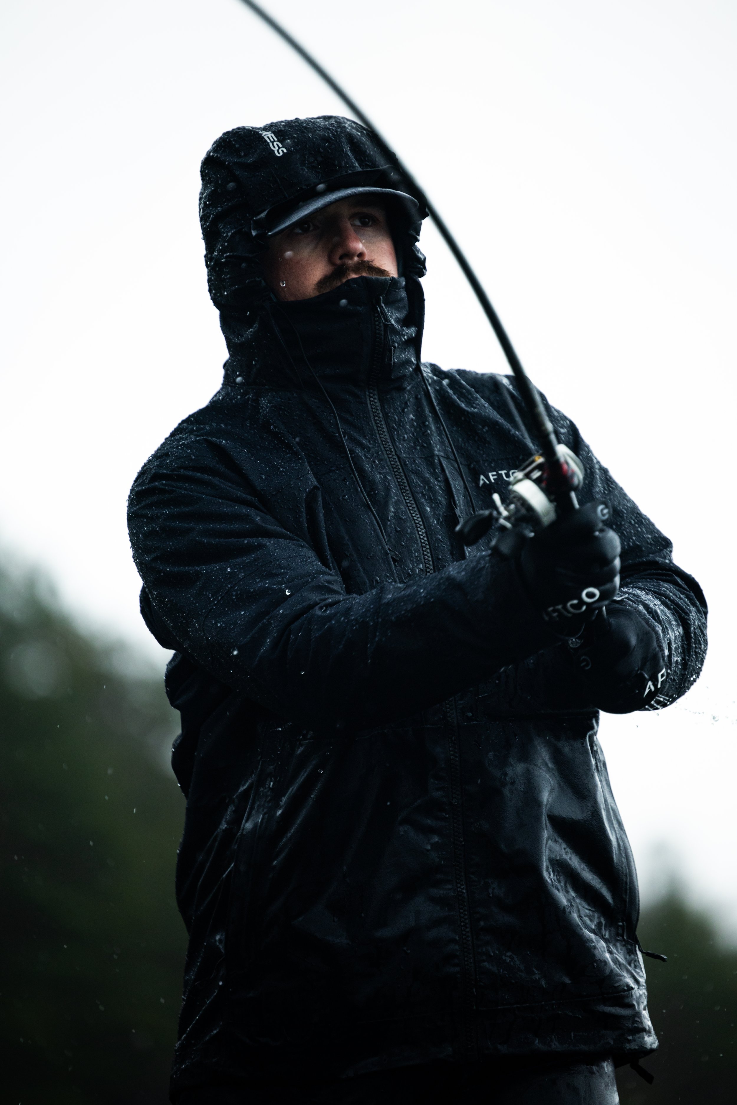 Man fishing in rainy weather, wearing a black rain jacket and hat, holding a fishing rod.