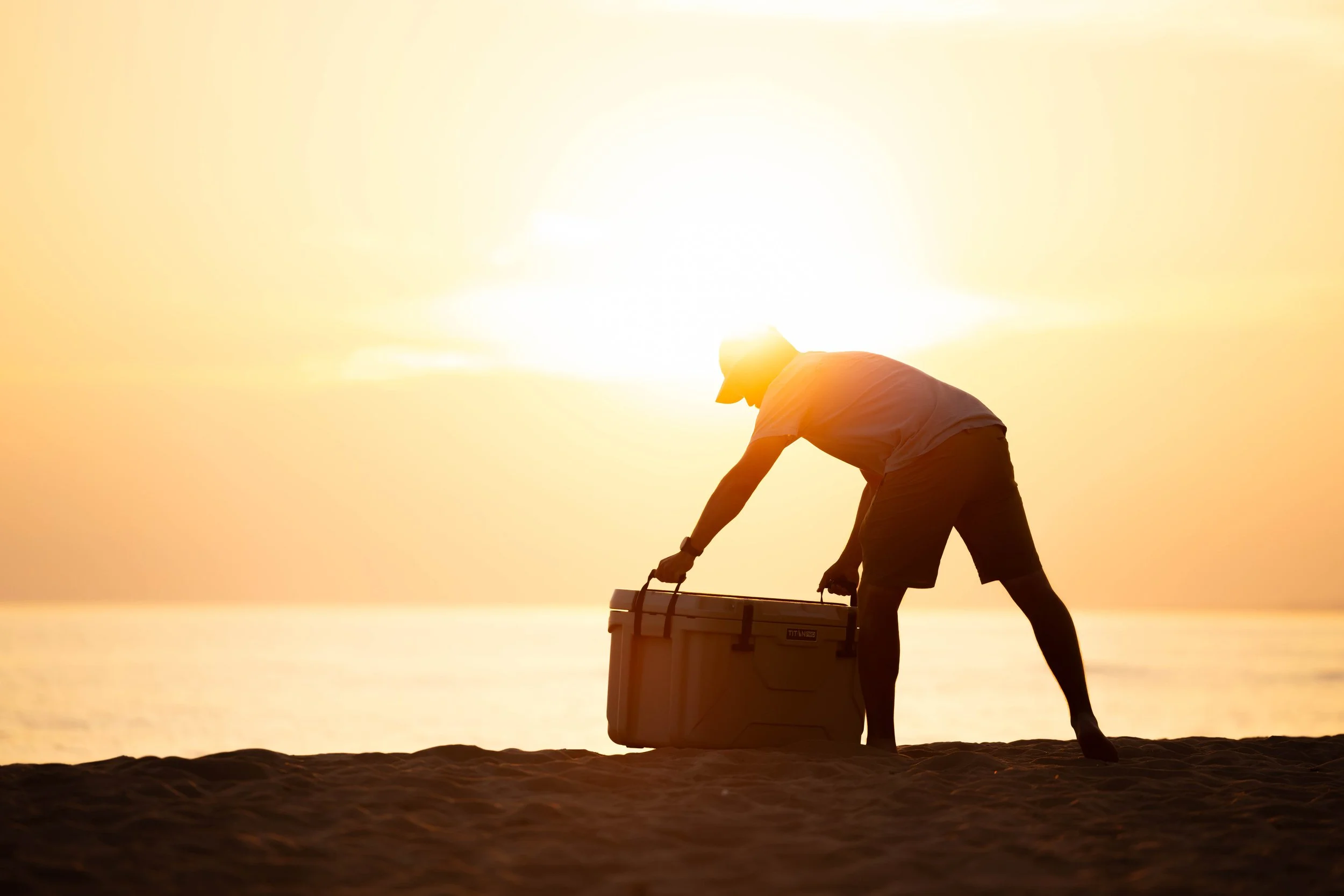 A person on the beach during sunset, lifting a cooler.