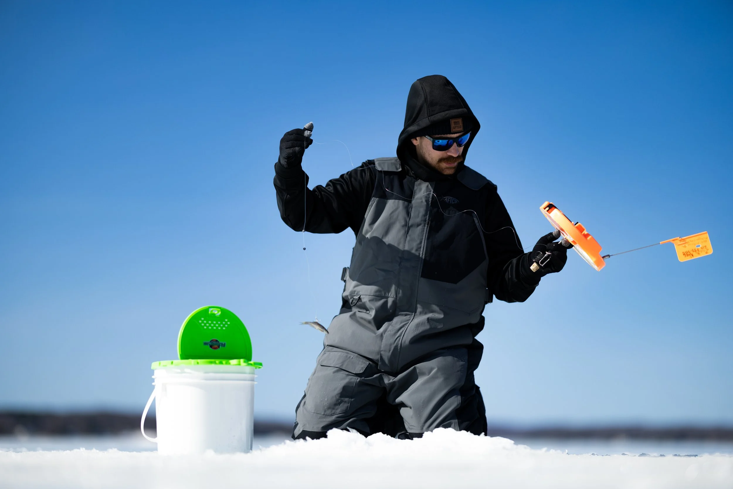 A man dressed in winter clothing, including a black hoodie, sunglasses, and gloves, is ice fishing on a clear day with a blue sky. He is holding a fishing line and a fishing device, with a plastic bucket nearby.