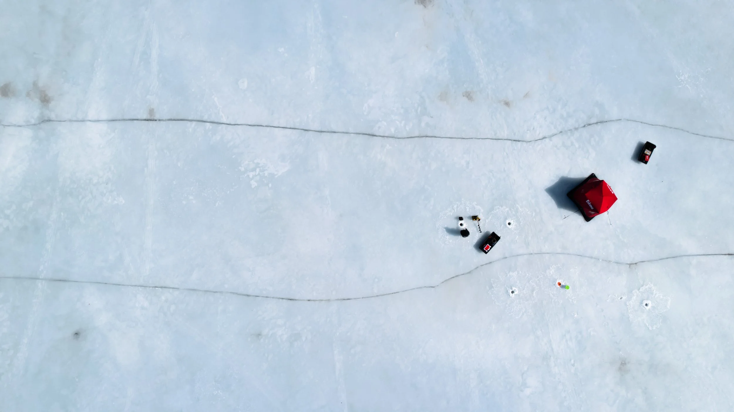 Aerial view of a snowy landscape with scattered equipment and a red tent, and tire tracks on the snow.
