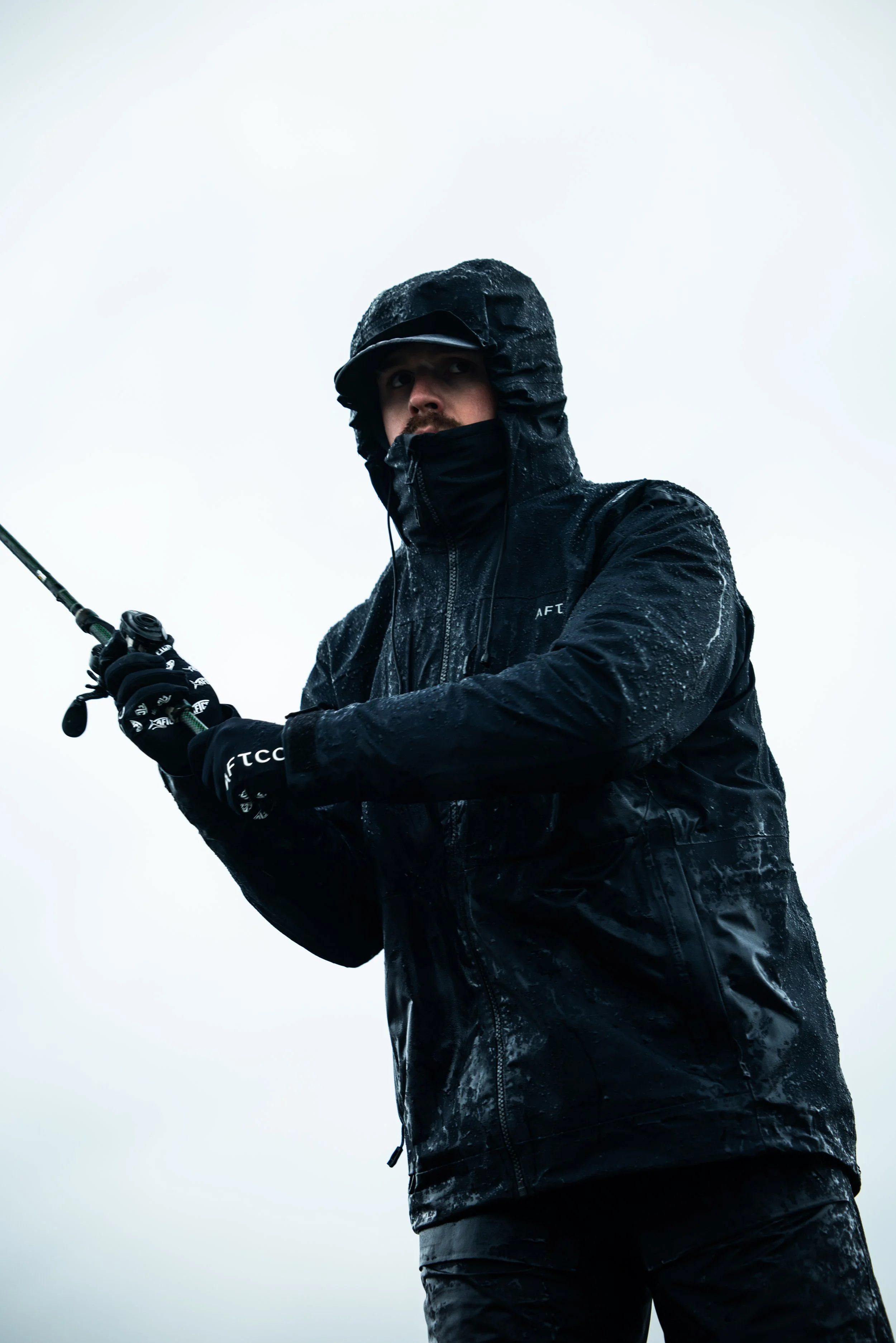 Person wearing a black waterproof jacket, gloves, and a hood, holding a fishing rod in an overcast outdoor setting.